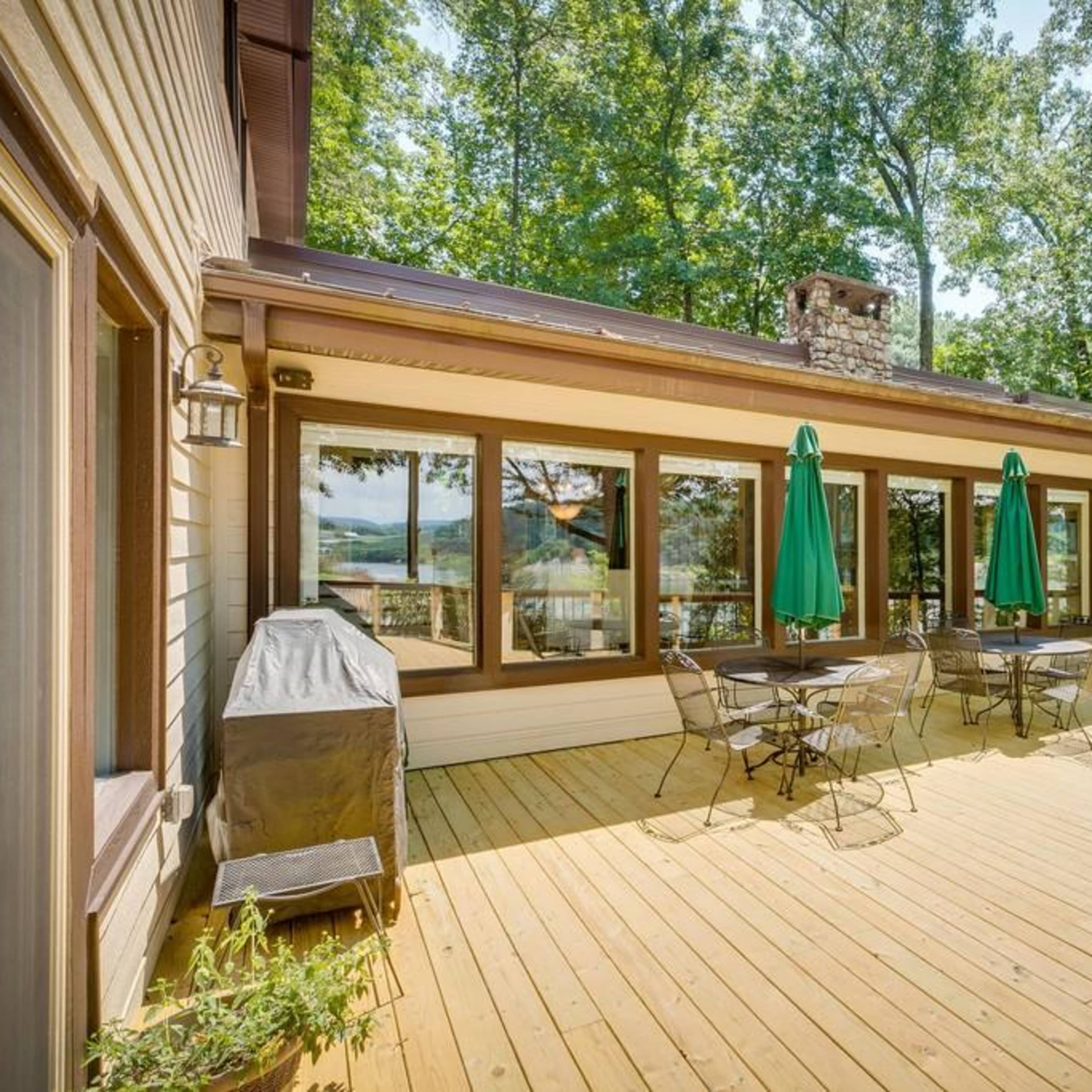 A wooden deck with outdoor furniture, green umbrellas, and large windows, surrounded by trees.