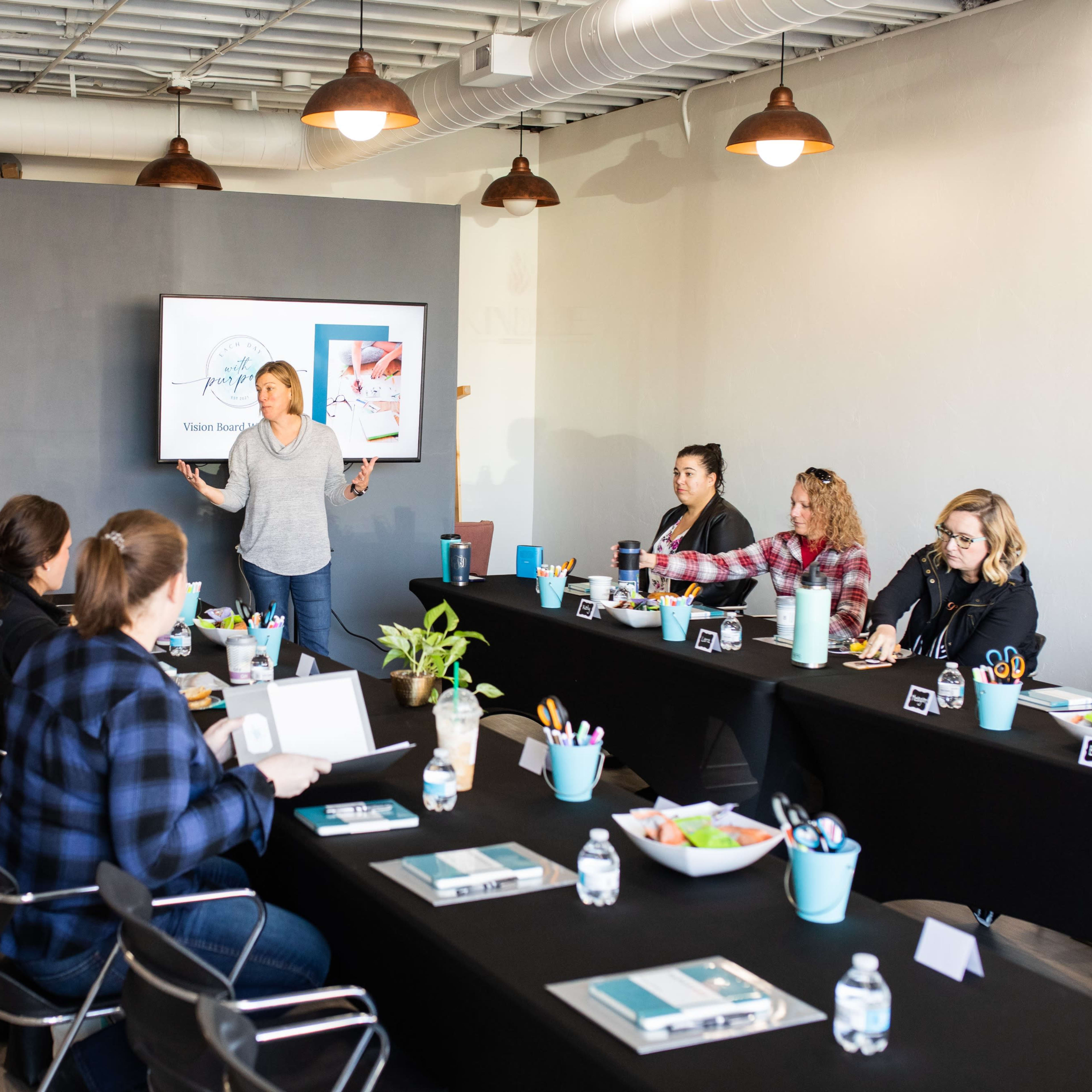 A group of people is seated at a long table in a meeting room, while a woman stands at the front presenting on a screen.