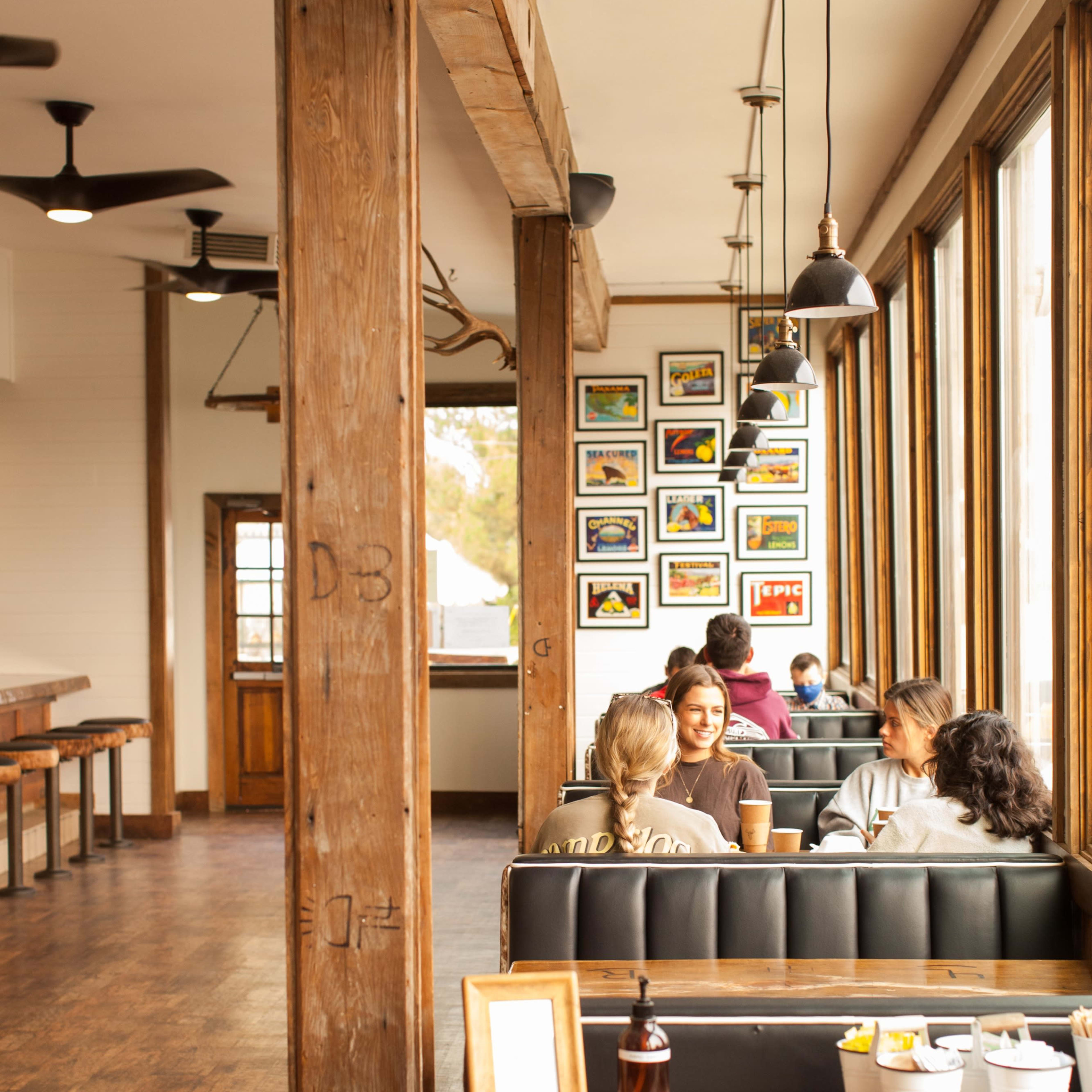A cozy café interior with wooden beams, tables, and patrons seated in booths enjoying their drinks, surrounded by framed artwork on the walls.