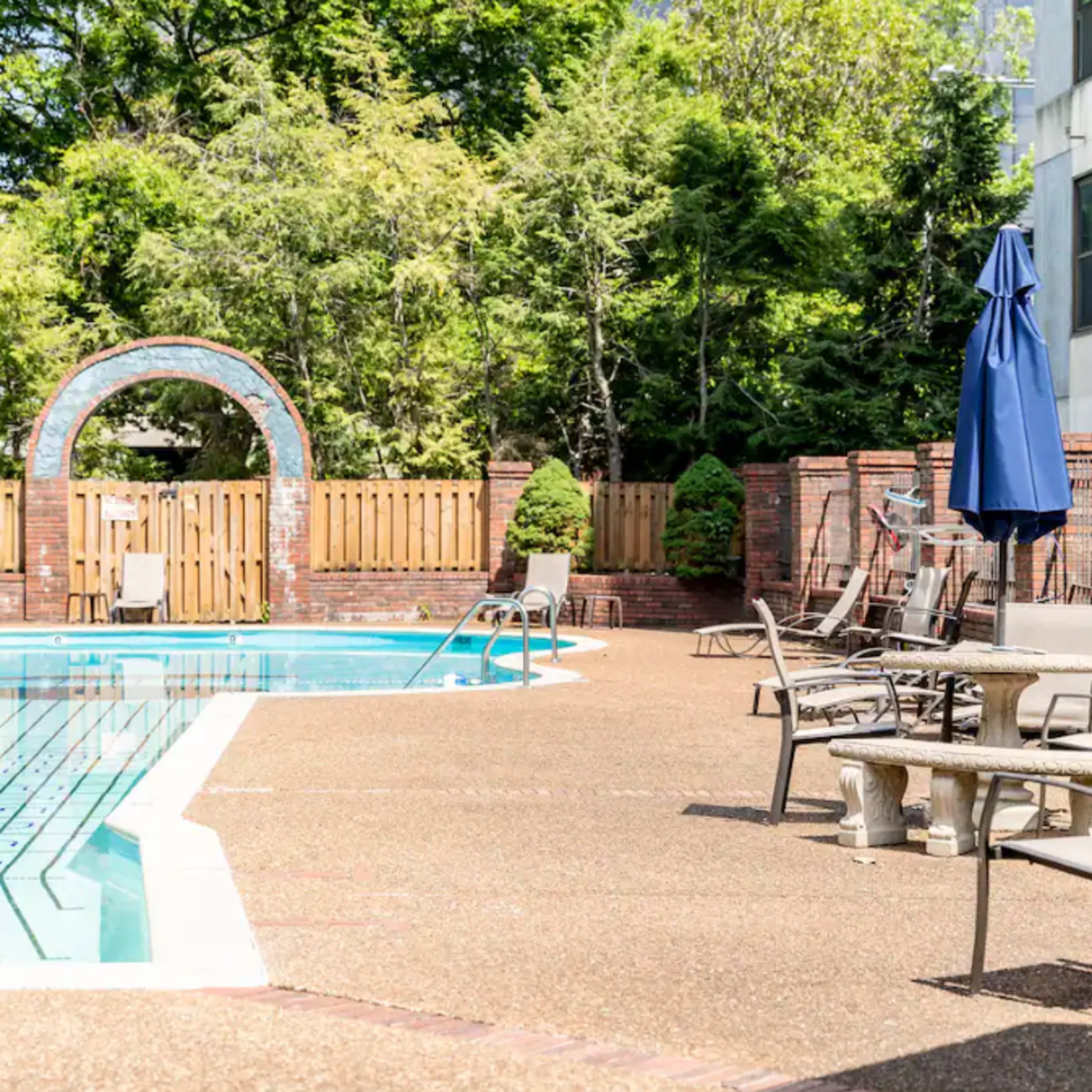 A swimming pool is surrounded by patio furniture and a wooden fence, with an archway in the background and greenery nearby.