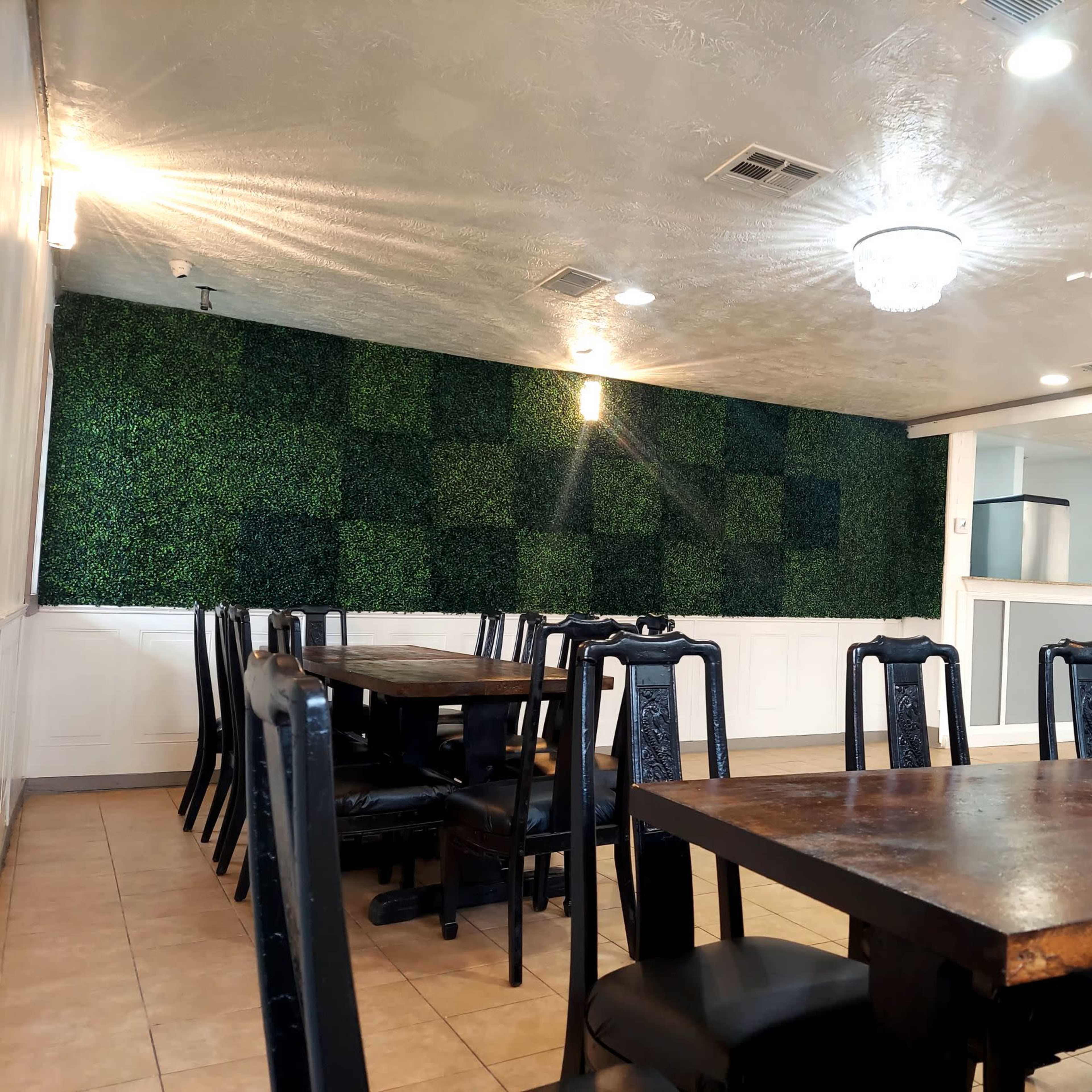 A dining area with black chairs and wooden tables, featuring a green wall of faux plants in the background.