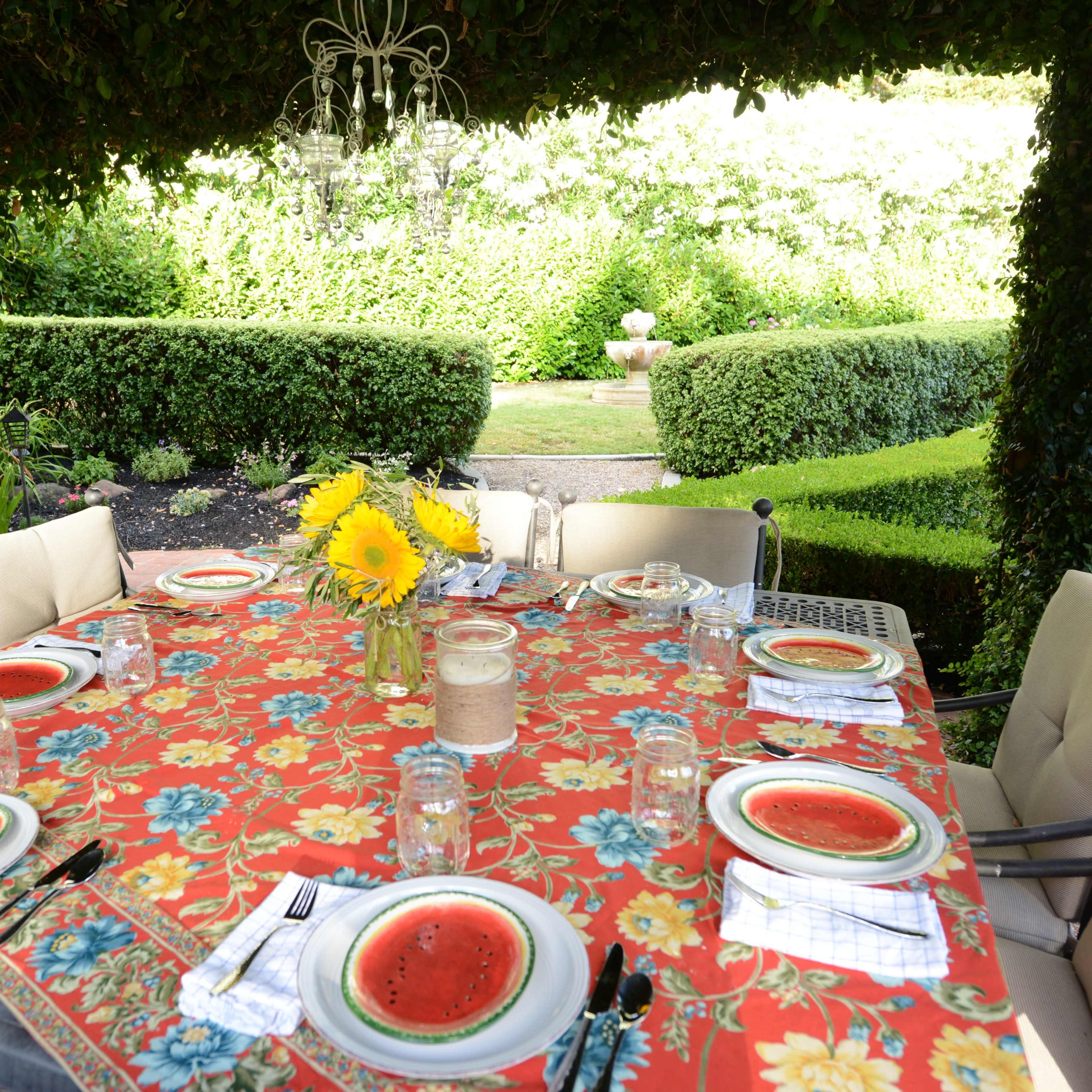 A neatly arranged dining table with plates of watermelon slices sits under a canopy of greenery, surrounded by a well-manicured garden.
