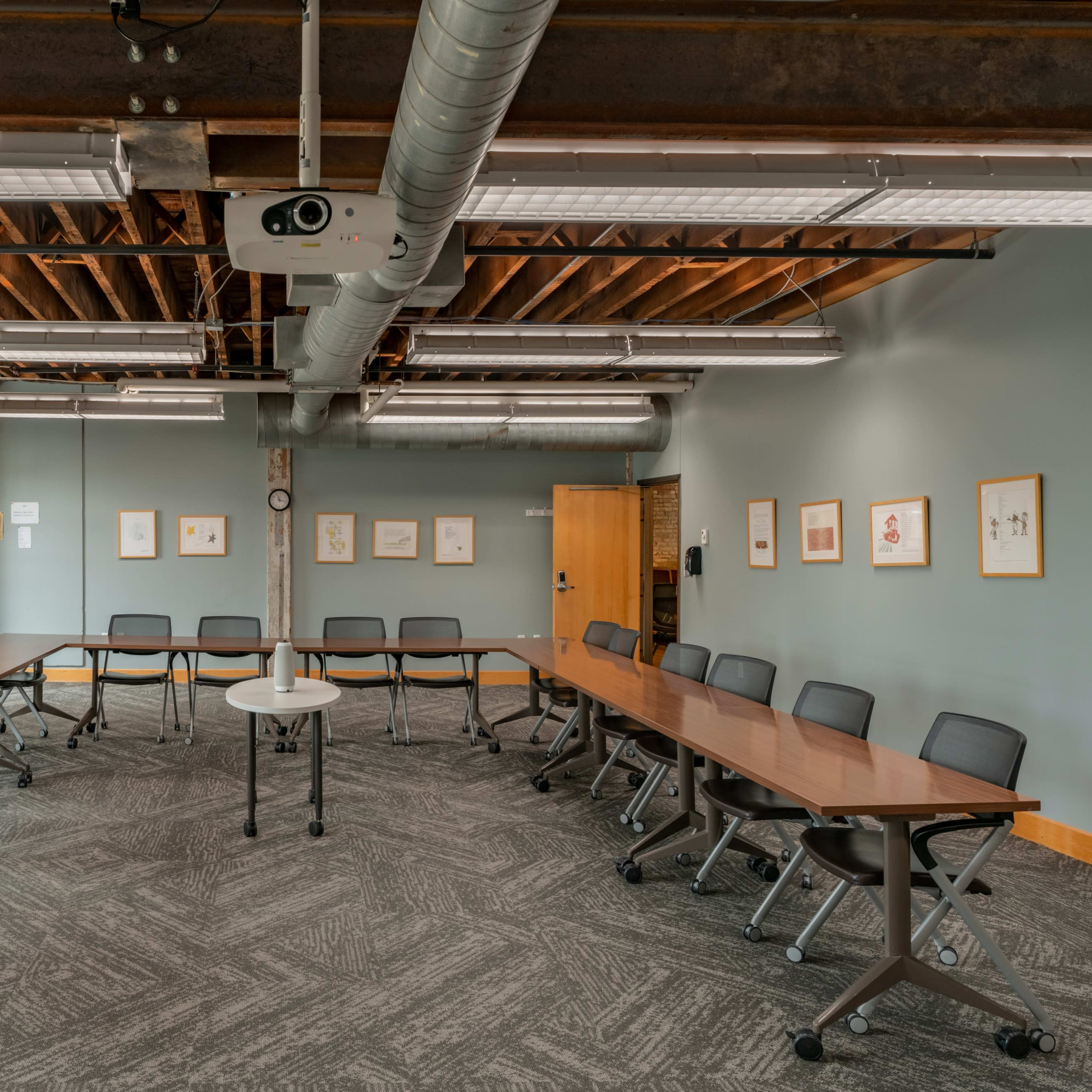 A spacious meeting room features a U-shaped arrangement of tables and chairs with exposed brick walls and overhead lighting.