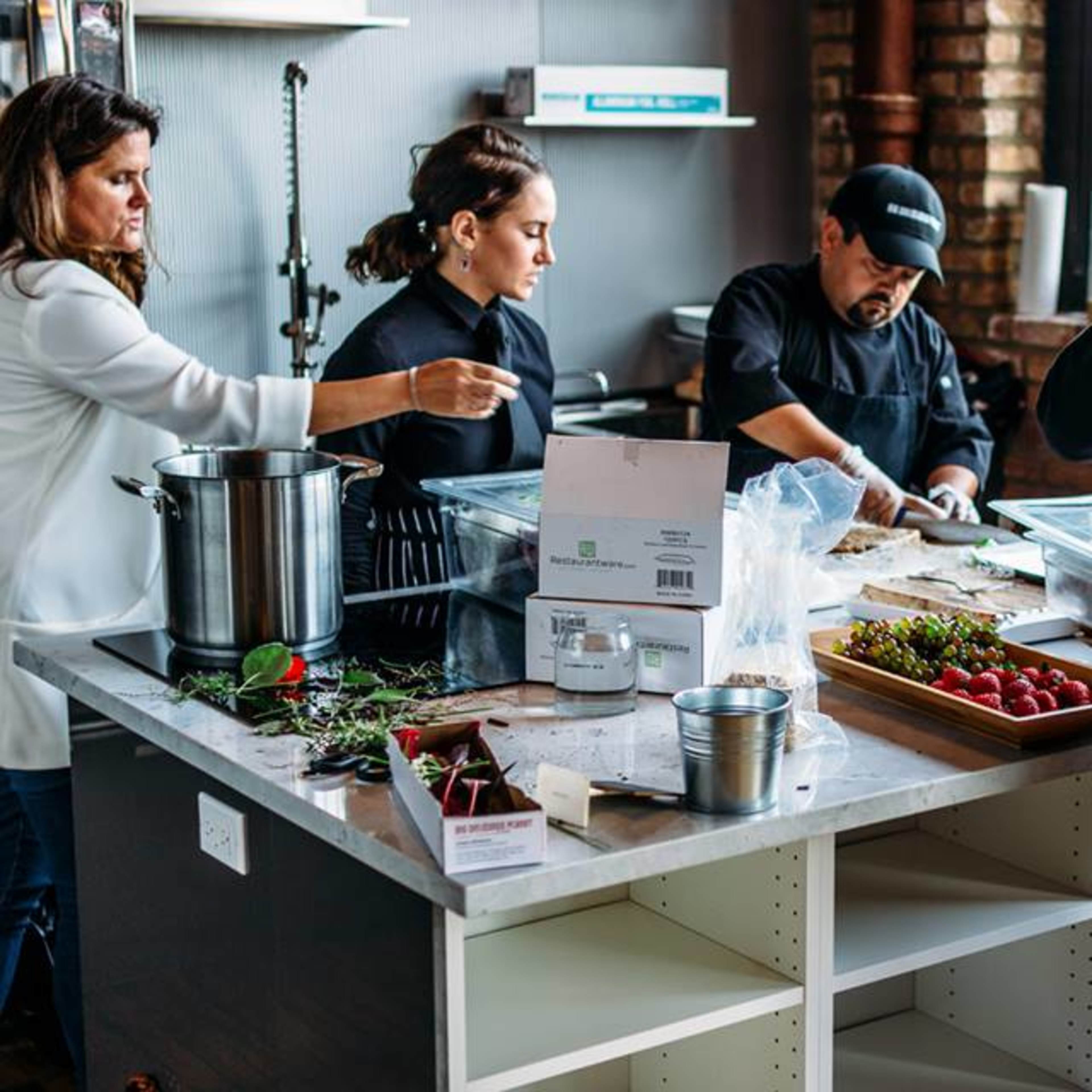 A group of chefs is preparing food at a kitchen counter with ingredients and cooking utensils arranged around them.