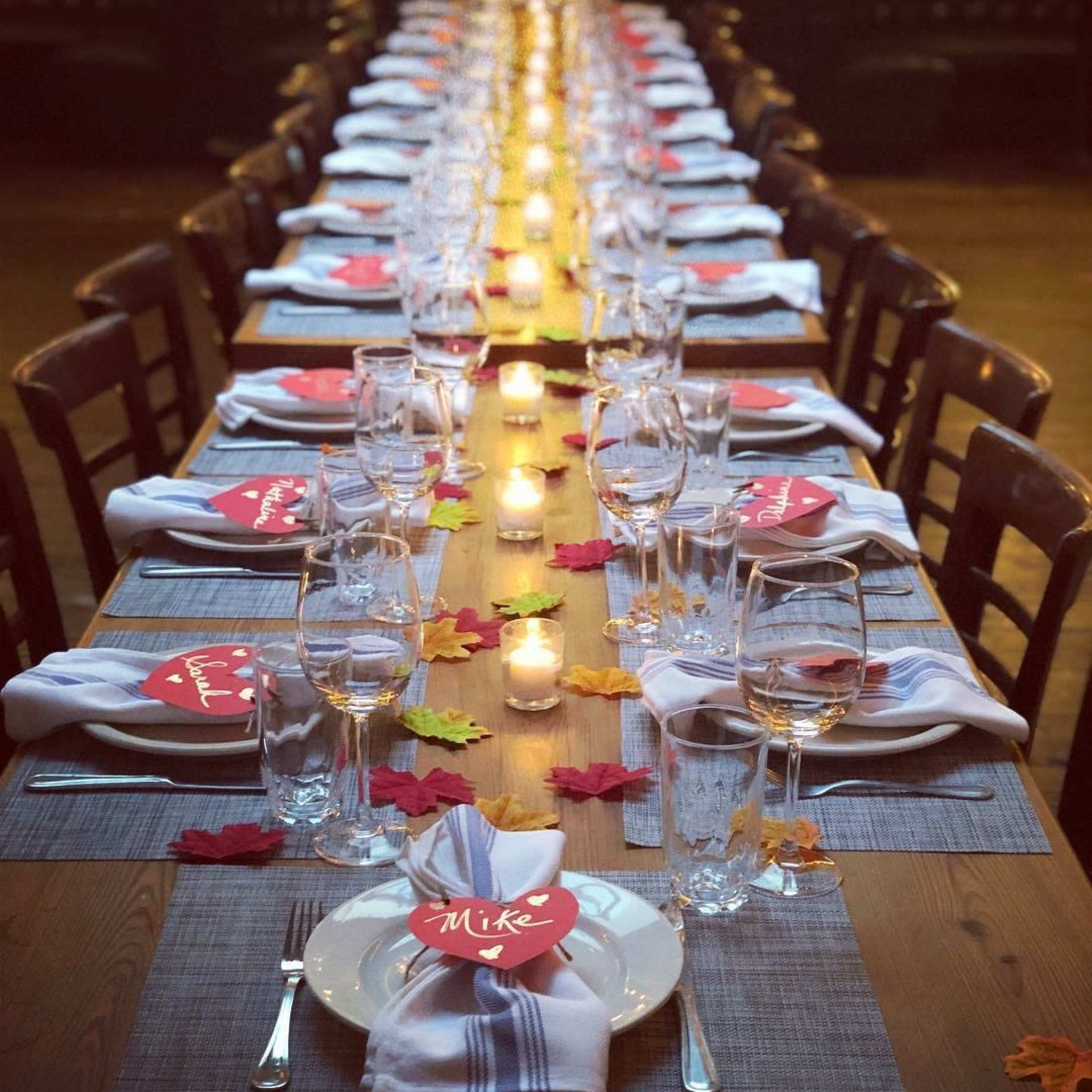 A long wooden table is set for a meal, featuring place cards, white napkins, and candlelight, with colorful leaves scattered decoratively across the table.
