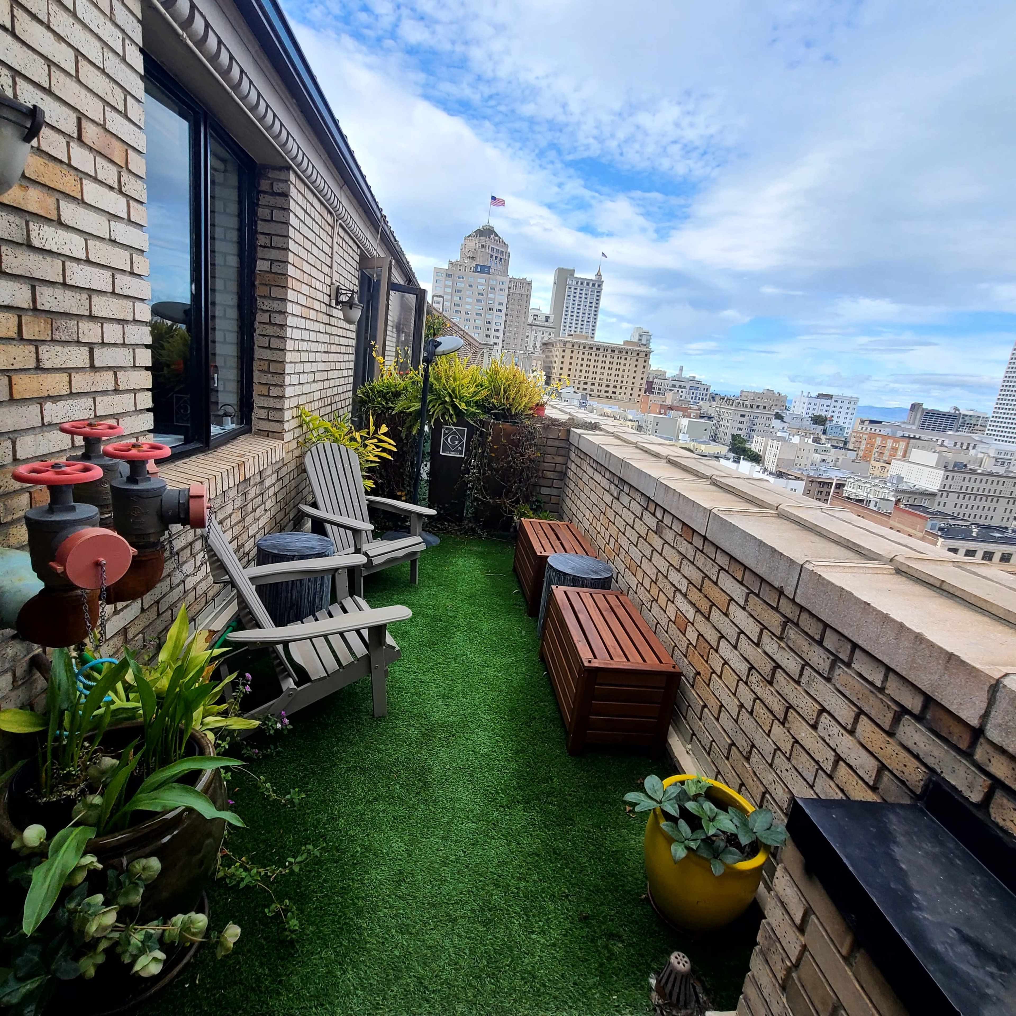 A rooftop balcony features green artificial turf, wooden benches, and potted plants, overlooking a city skyline under a partly cloudy sky.