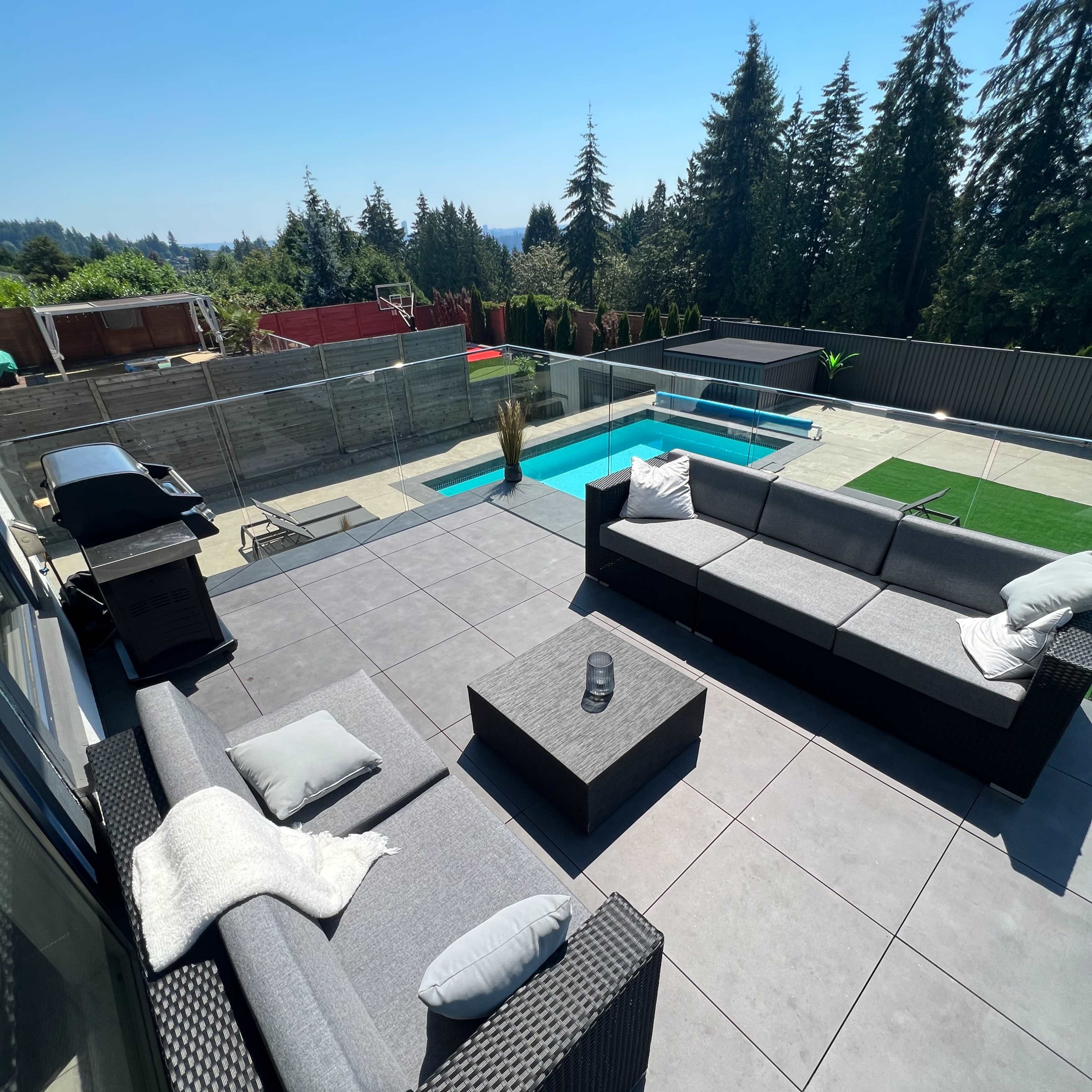 A modern outdoor patio area with a gray tiled floor, featuring two sectional sofas, a coffee table, a grill, and a swimming pool surrounded by trees.