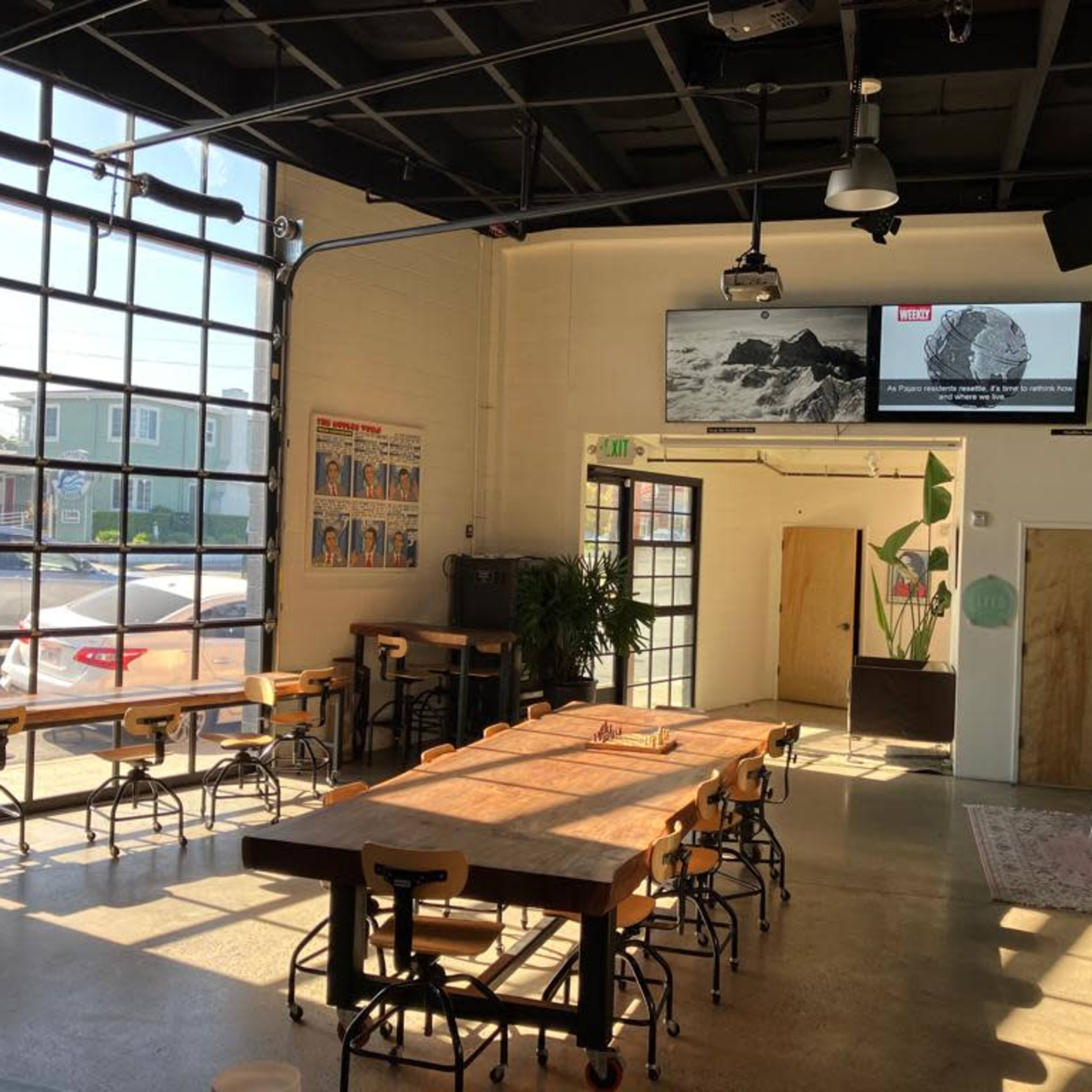 The interior of a spacious café features large windows, several wooden tables, and a screen displaying an image on the wall.