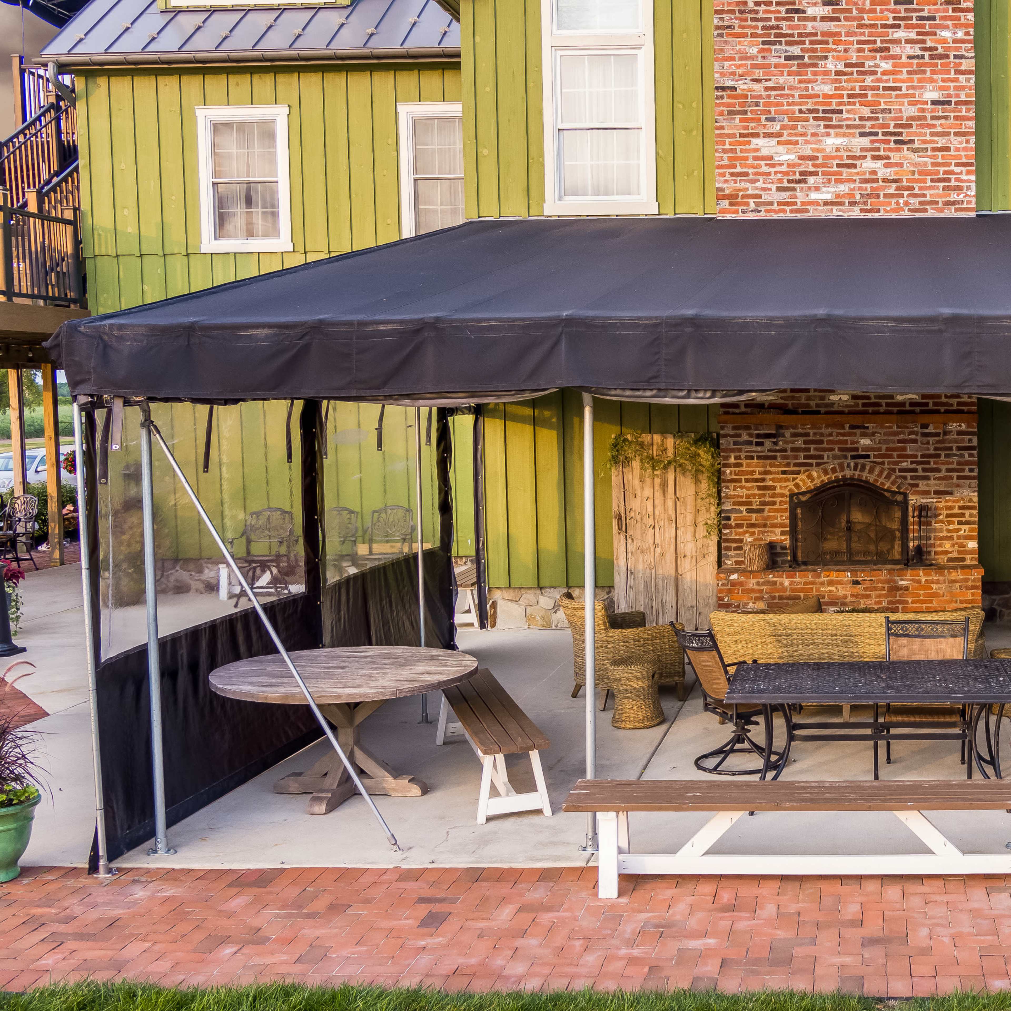 A large black tent covers a patio area with outdoor seating, a stone fireplace, and an adjacent wooden structure beside a green and brick building.