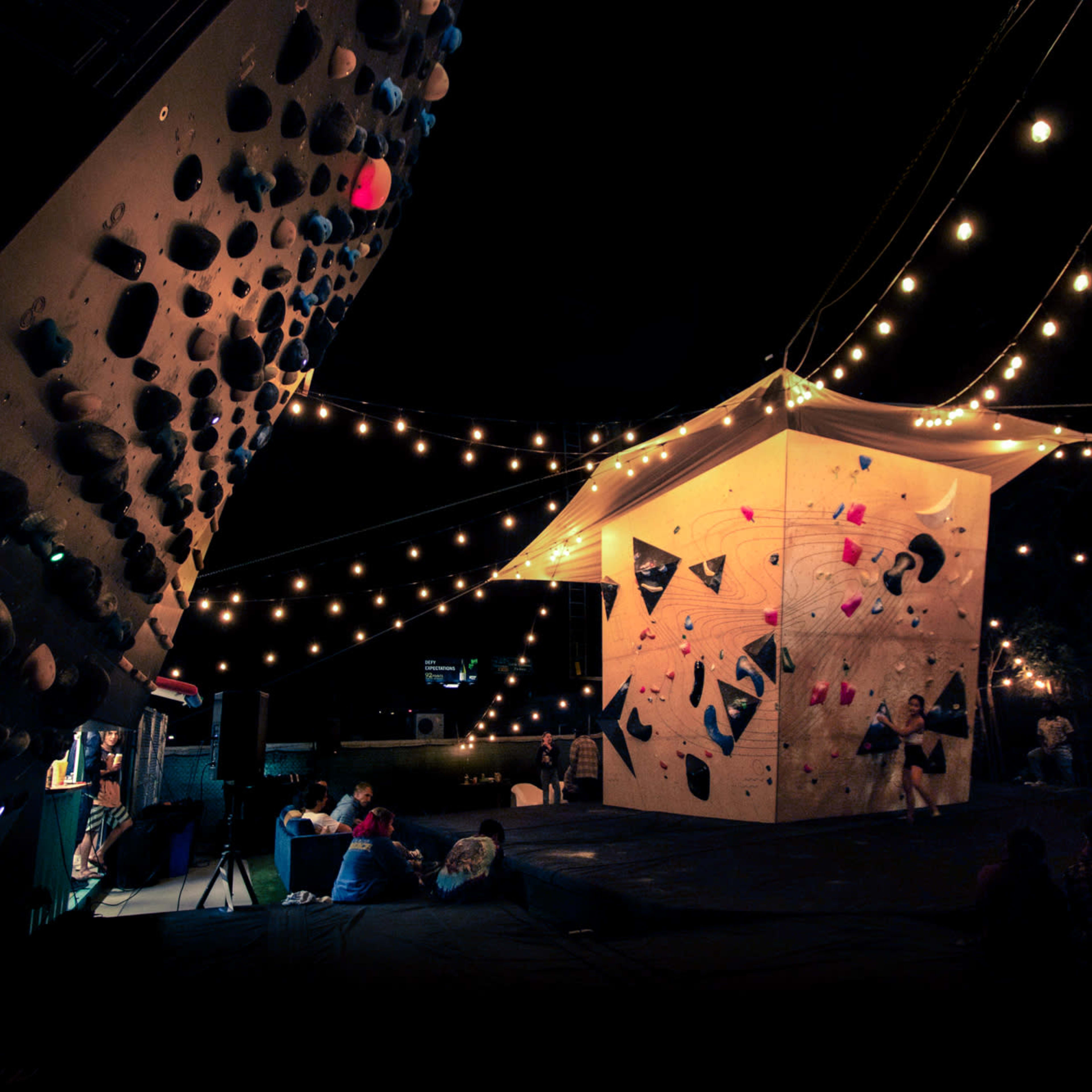 A climbing structure lit by string lights is set up outdoors, with people gathered nearby.