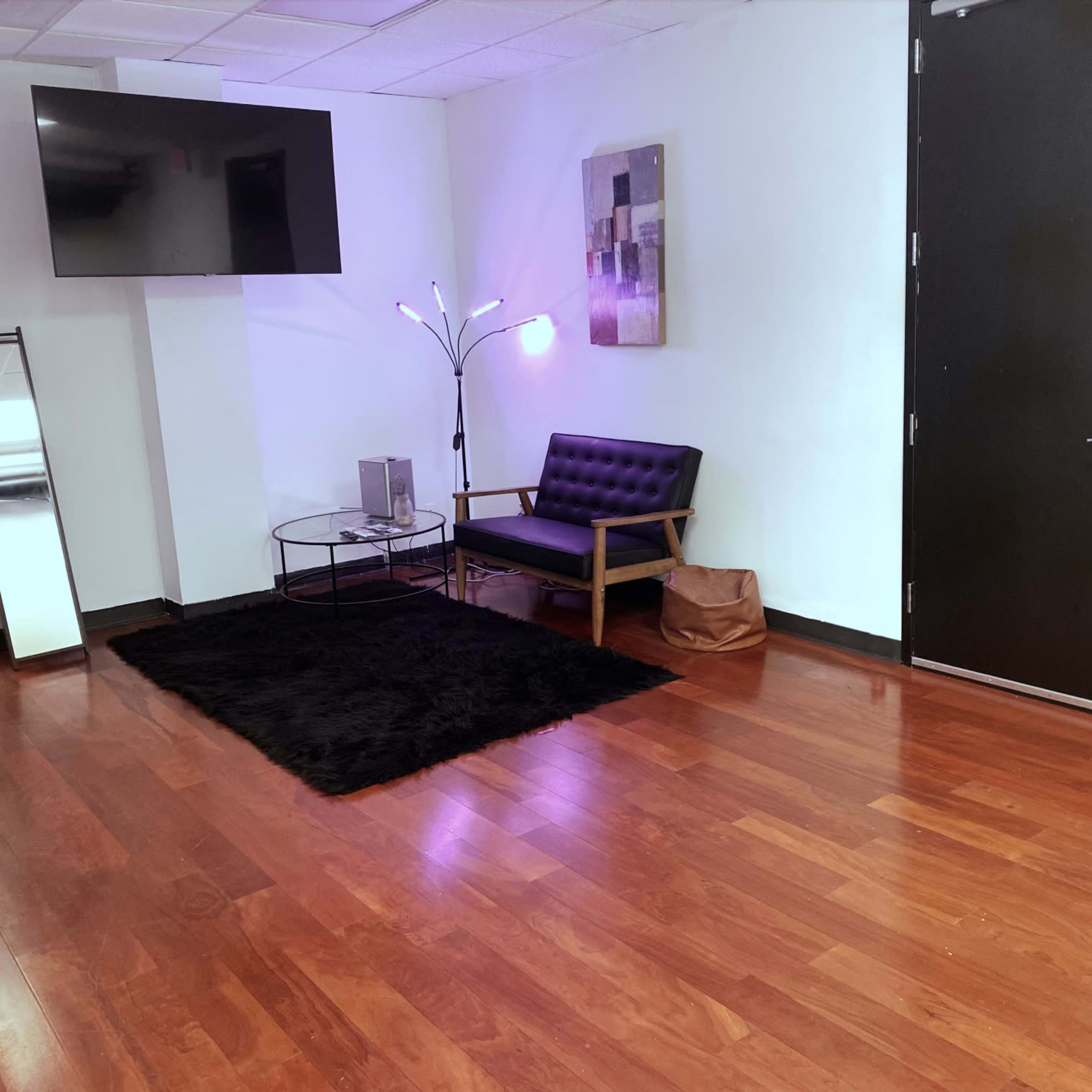 A minimalist interior with a black sofa, a floor lamp, a television mounted on the wall, and a mirrored closet, all set on wooden flooring.