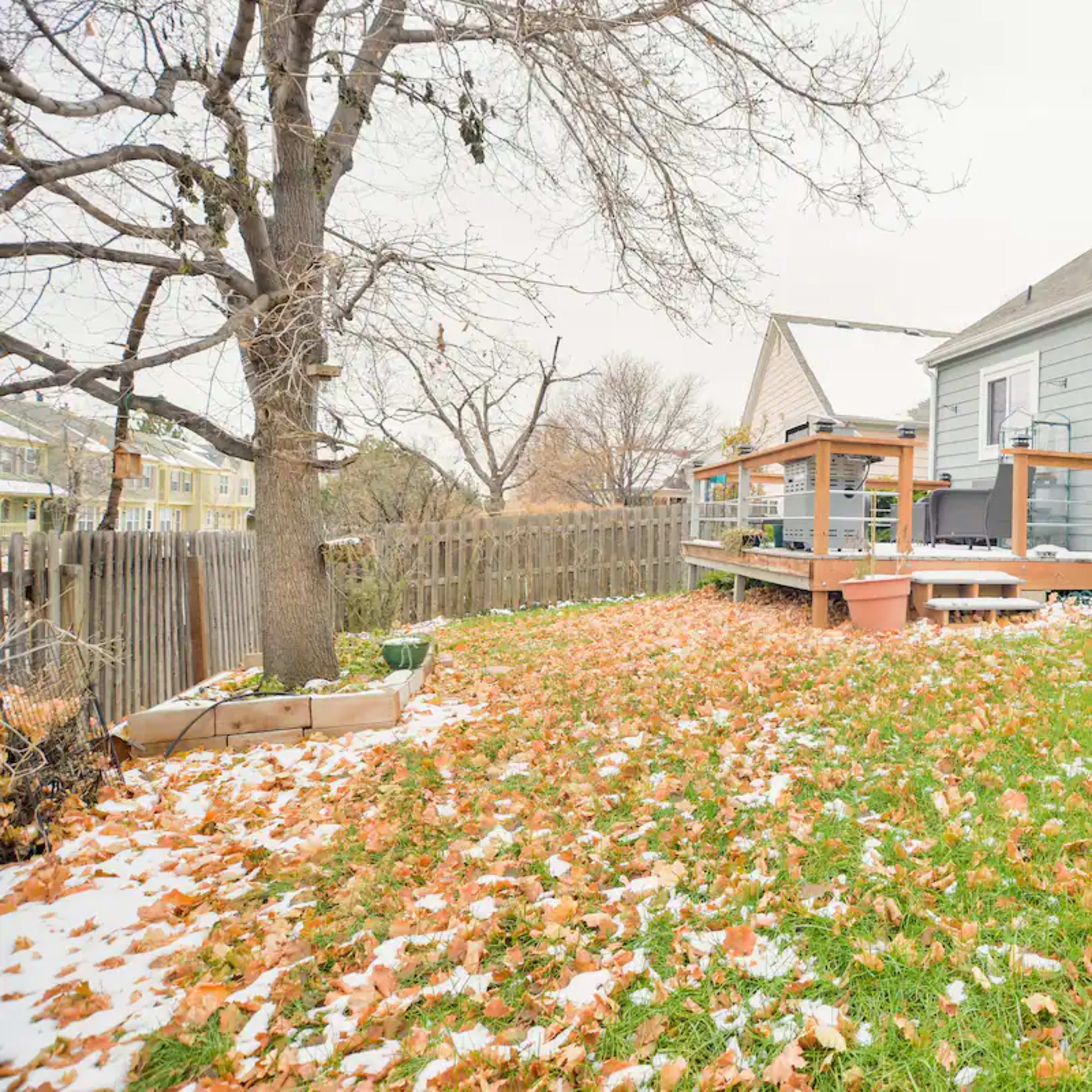 A backyard features a wooden deck surrounded by scattered leaves and patches of snow near a gray house.