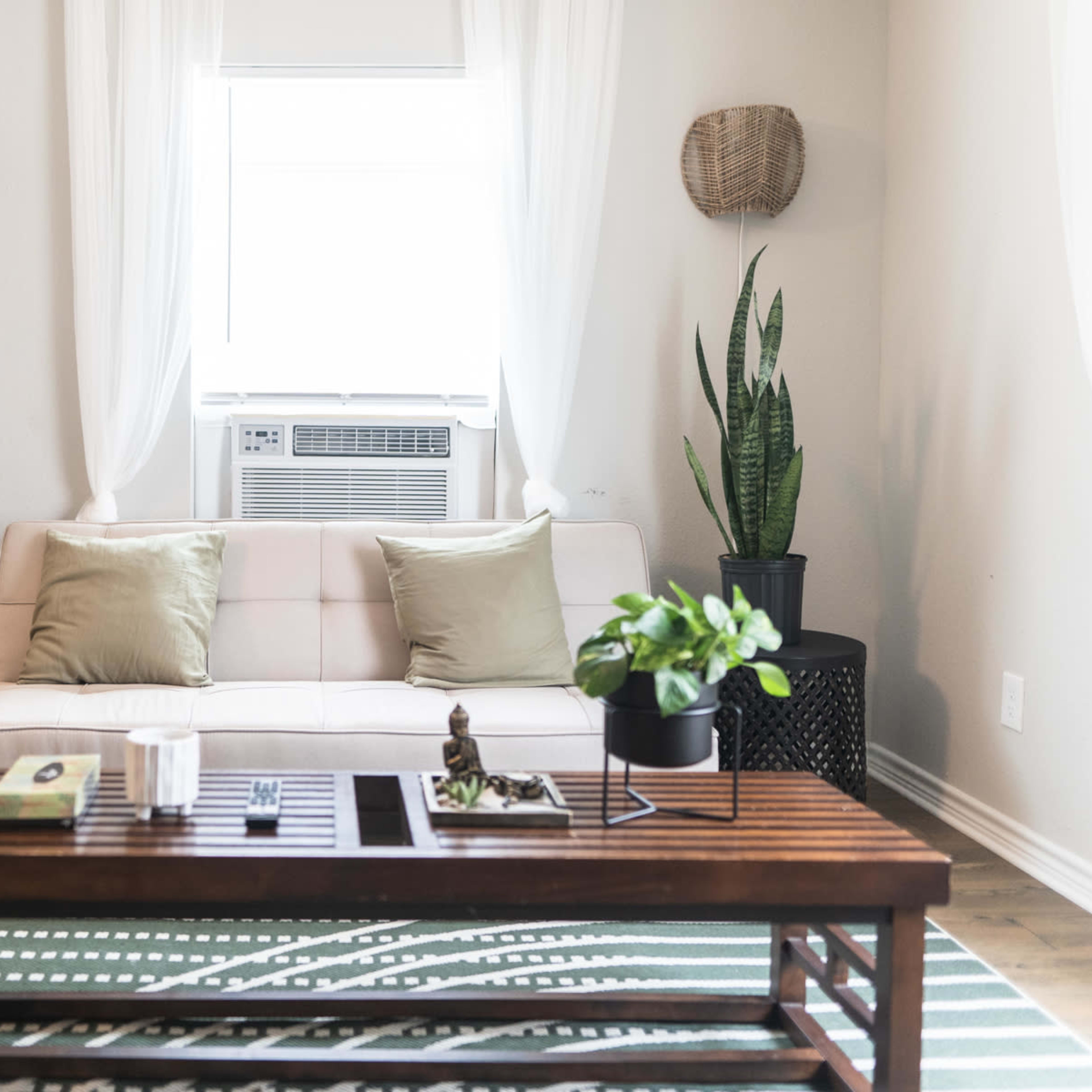 A minimalist living room features a beige sofa with two cushions, a wooden coffee table, and potted plants beside an air conditioning unit.