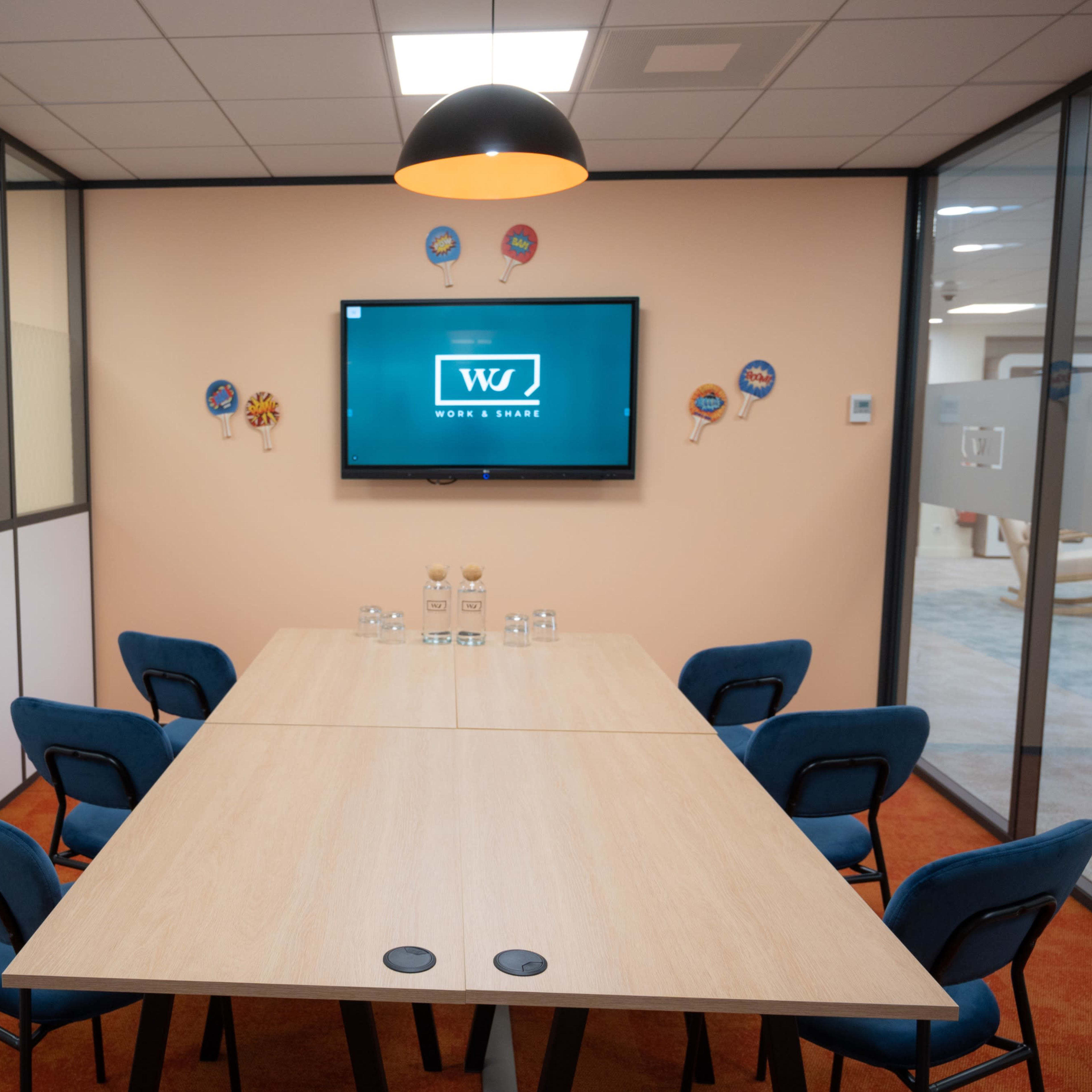 A modern conference room with a large wooden table, six blue chairs, and a wall-mounted television displaying a logo.