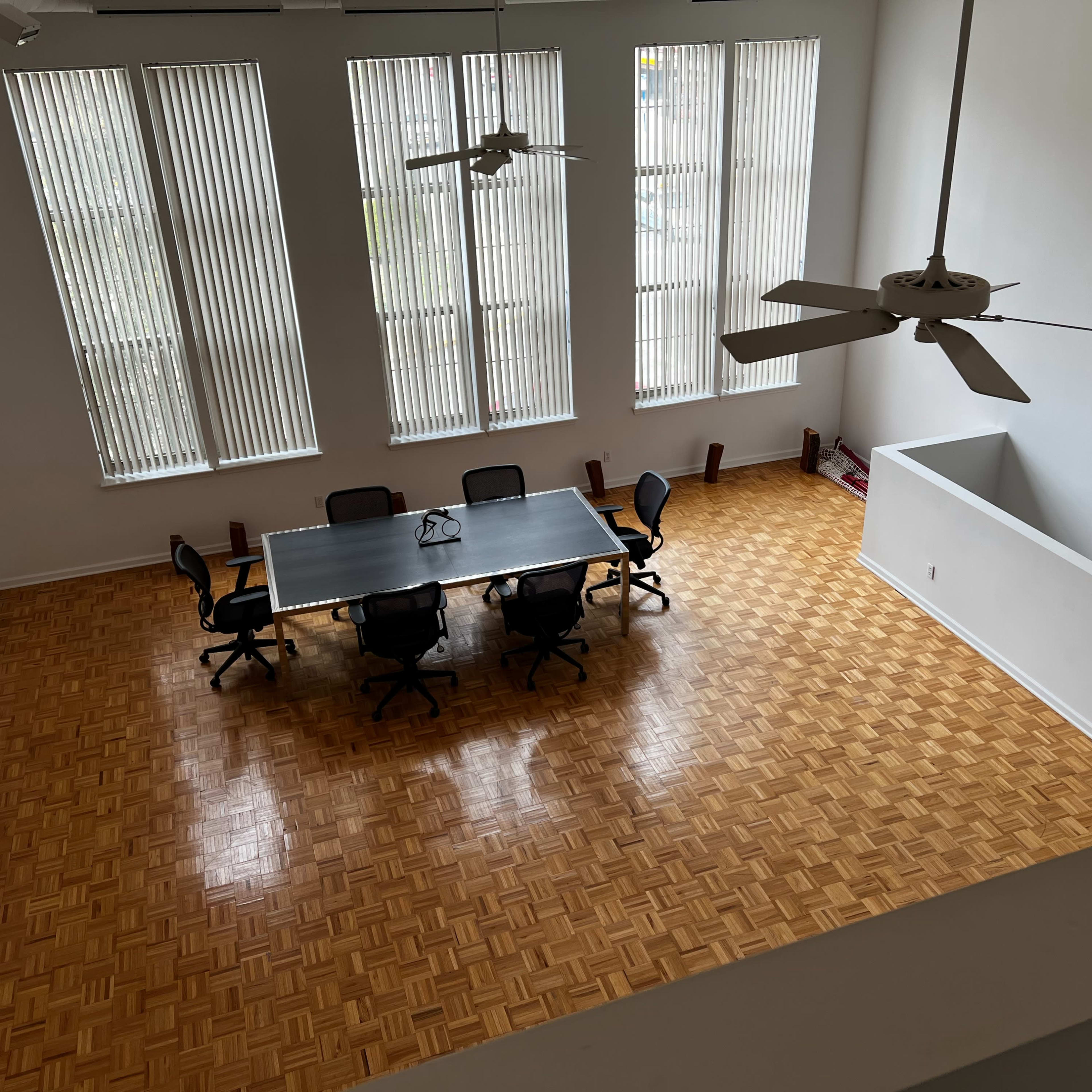 A spacious room with a large table surrounded by chairs, featuring tall windows and a wooden floor.