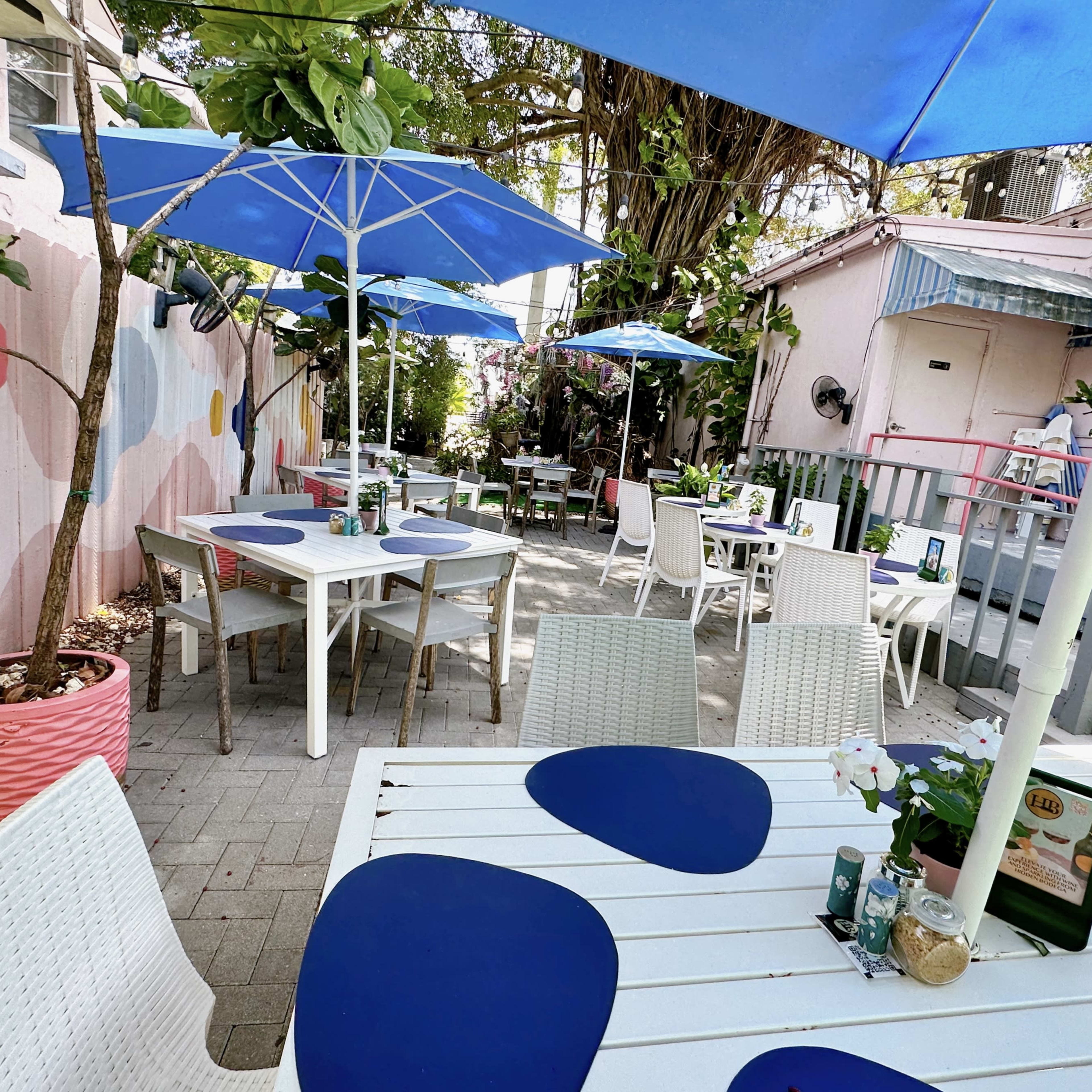 A patio dining area features white tables with blue placemats, surrounded by potted plants and umbrellas.