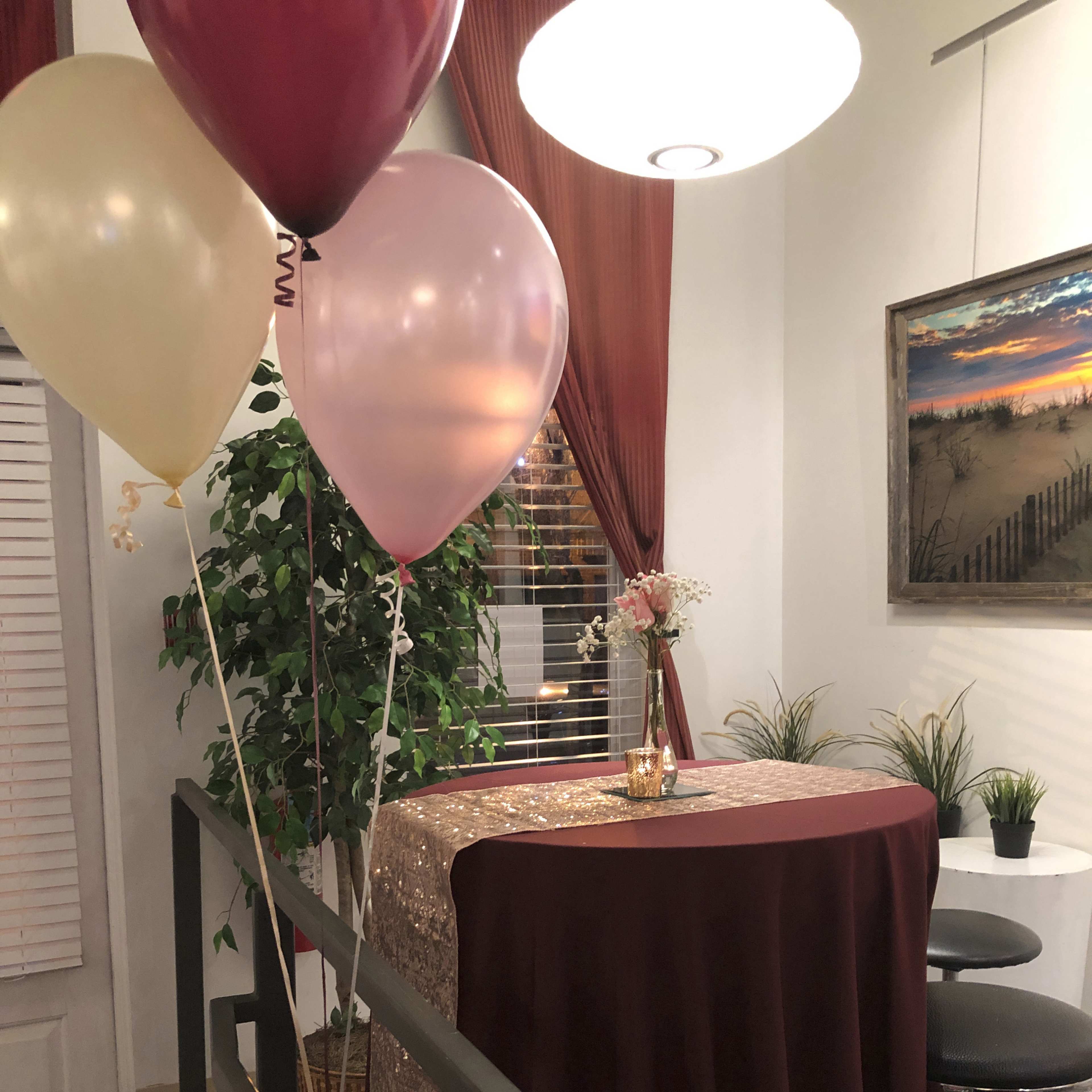 A small indoor space features a tall table draped in a maroon tablecloth, surrounded by balloons and a potted plant, with a painting of a beach scene on the wall.