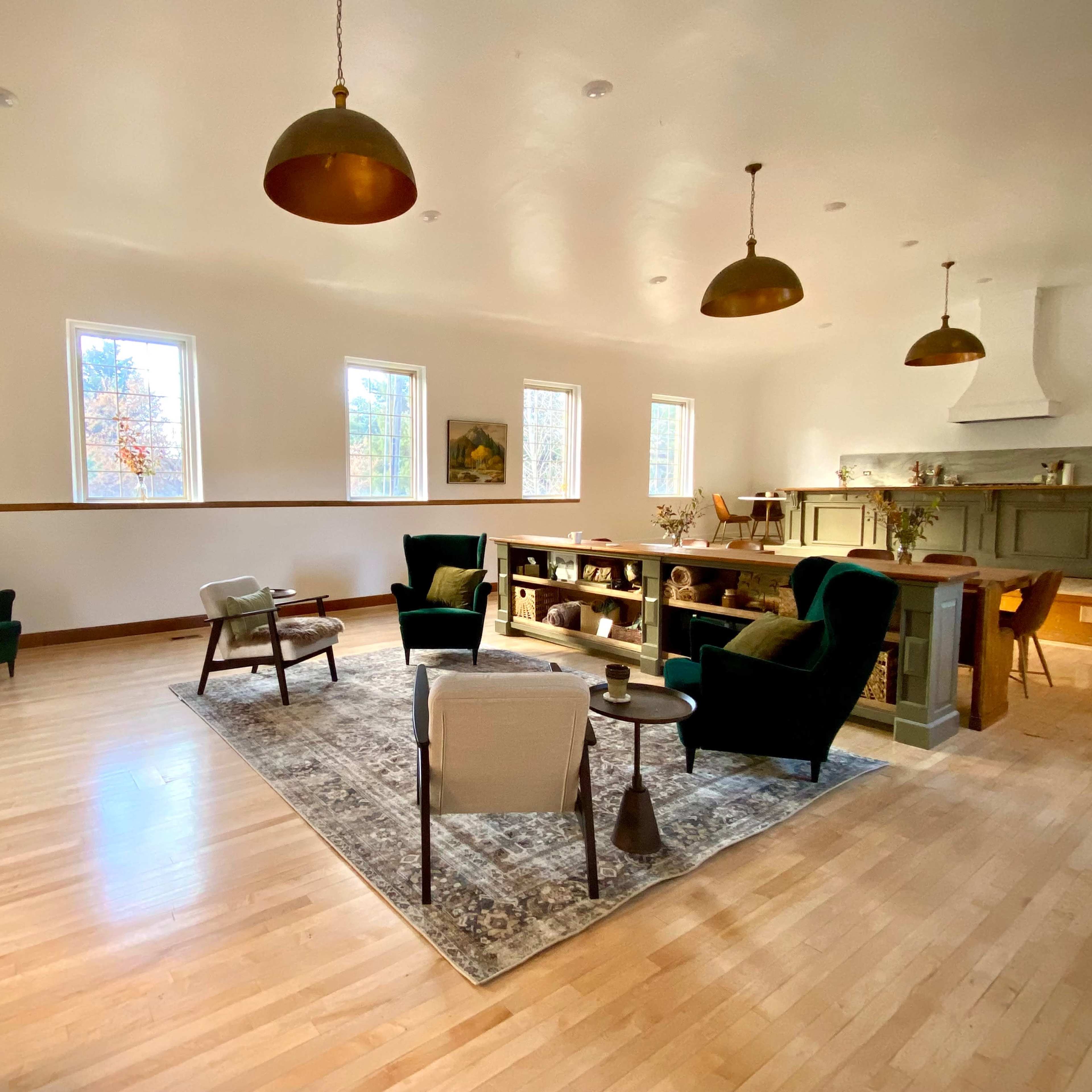 The spacious room features green chairs, a coffee table, a rug, and large windows that bring in natural light.