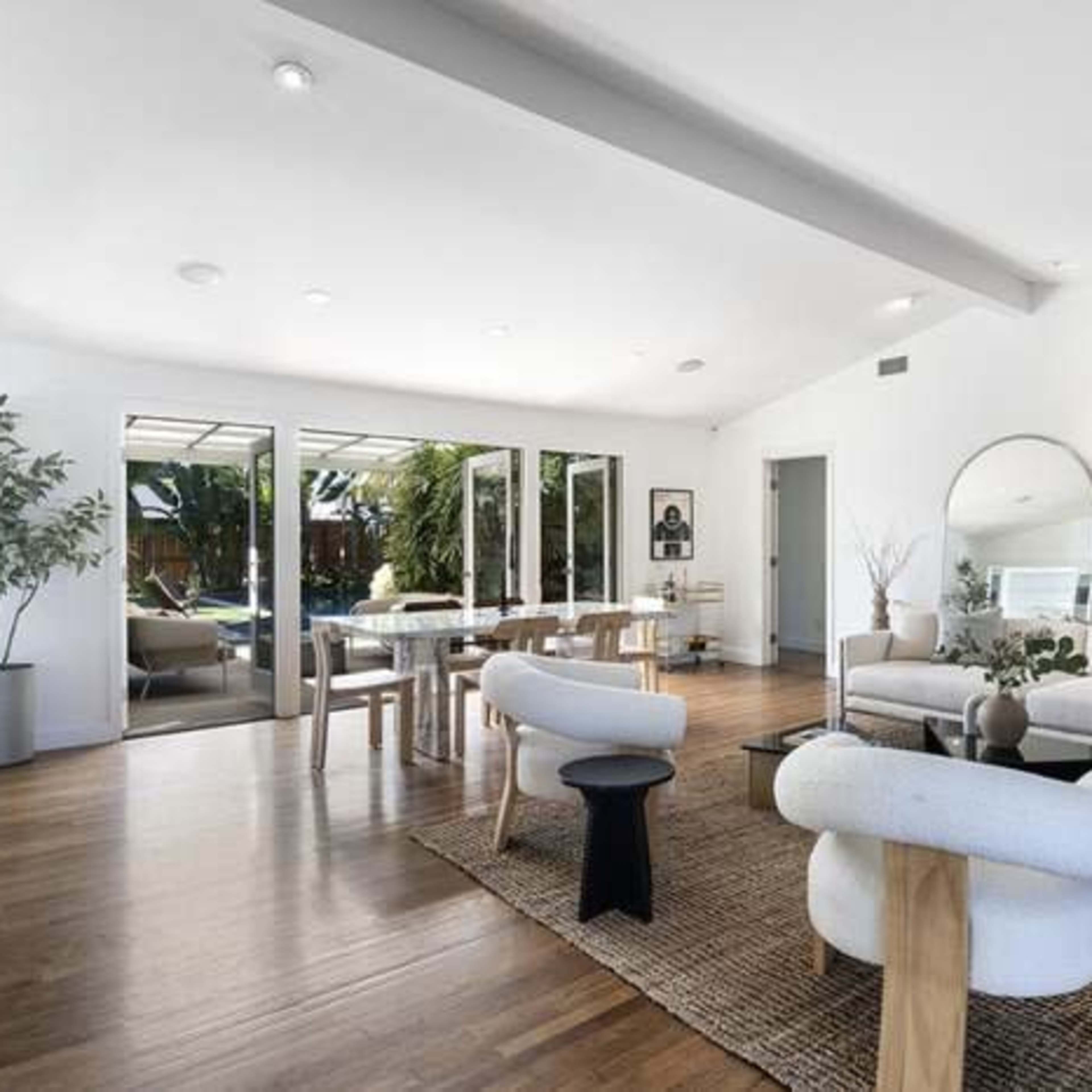 A modern living space with a light-colored sofa, wooden dining table, and large windows looking out to a garden.