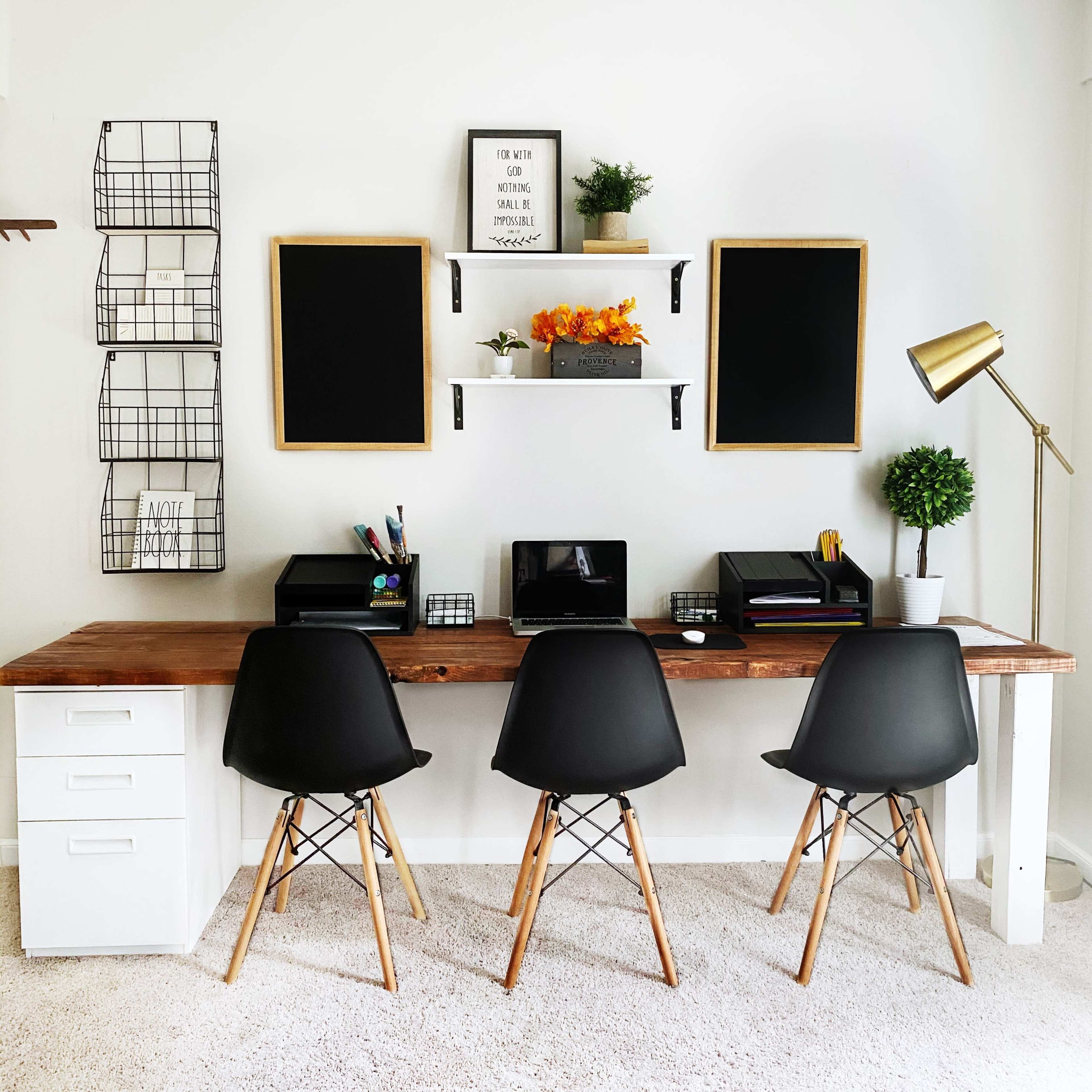 A modern workspace features a long wooden desk with three black chairs, storage bins, and decorative elements on the wall.