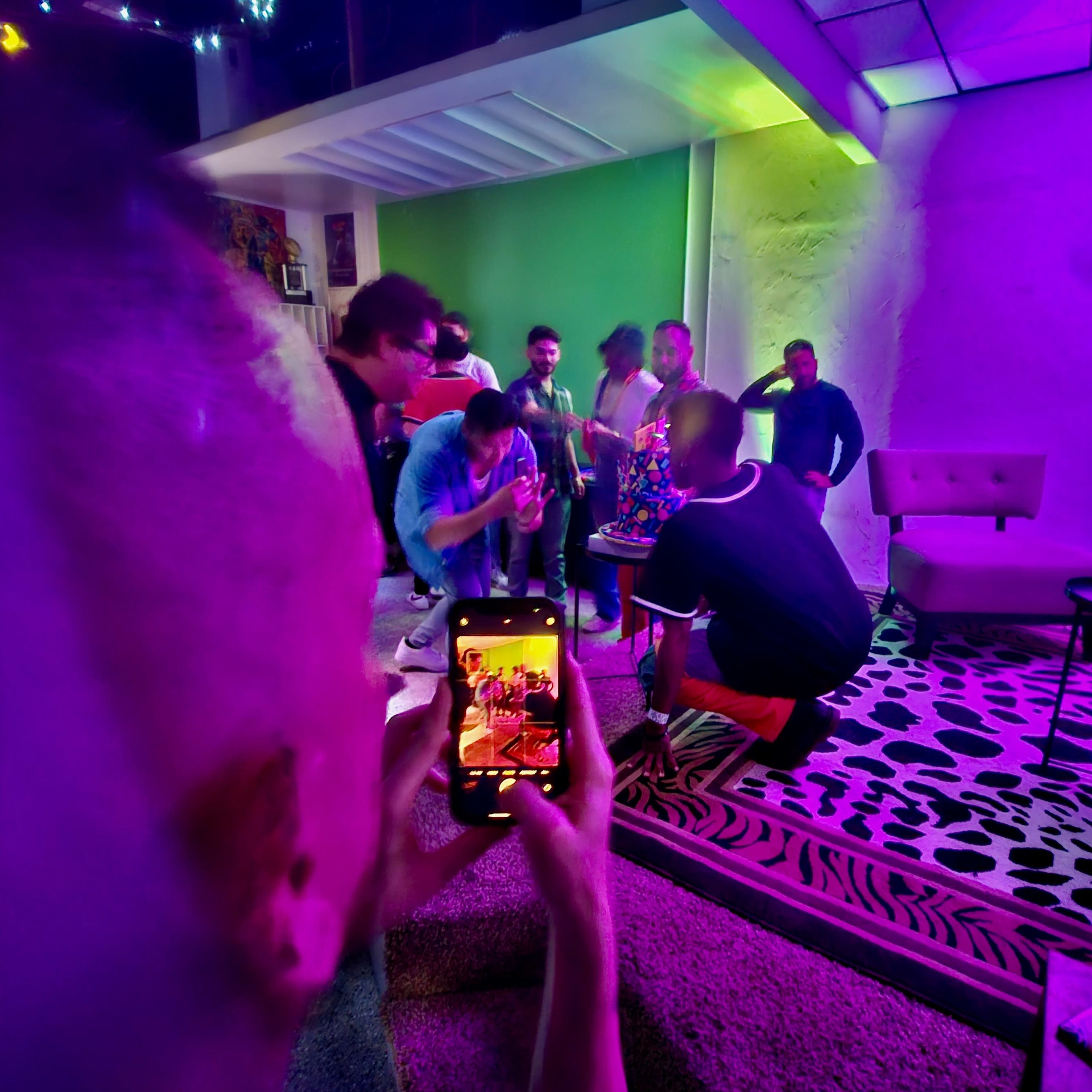 A group of people gathers in a brightly lit room, some dancing while one person records the scene on a smartphone.