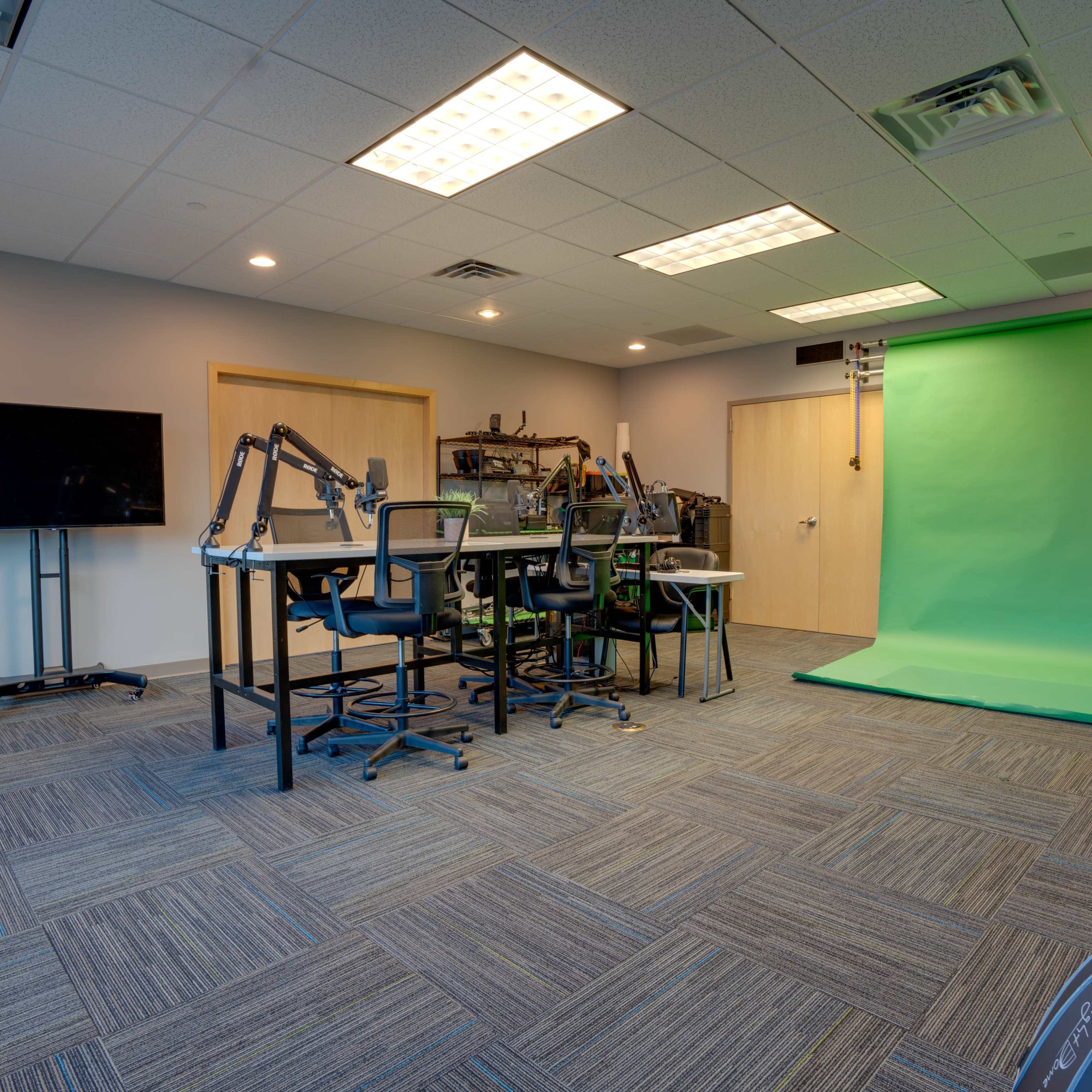 A video production studio featuring a green screen, several workstations with chairs, and a television on a stand.