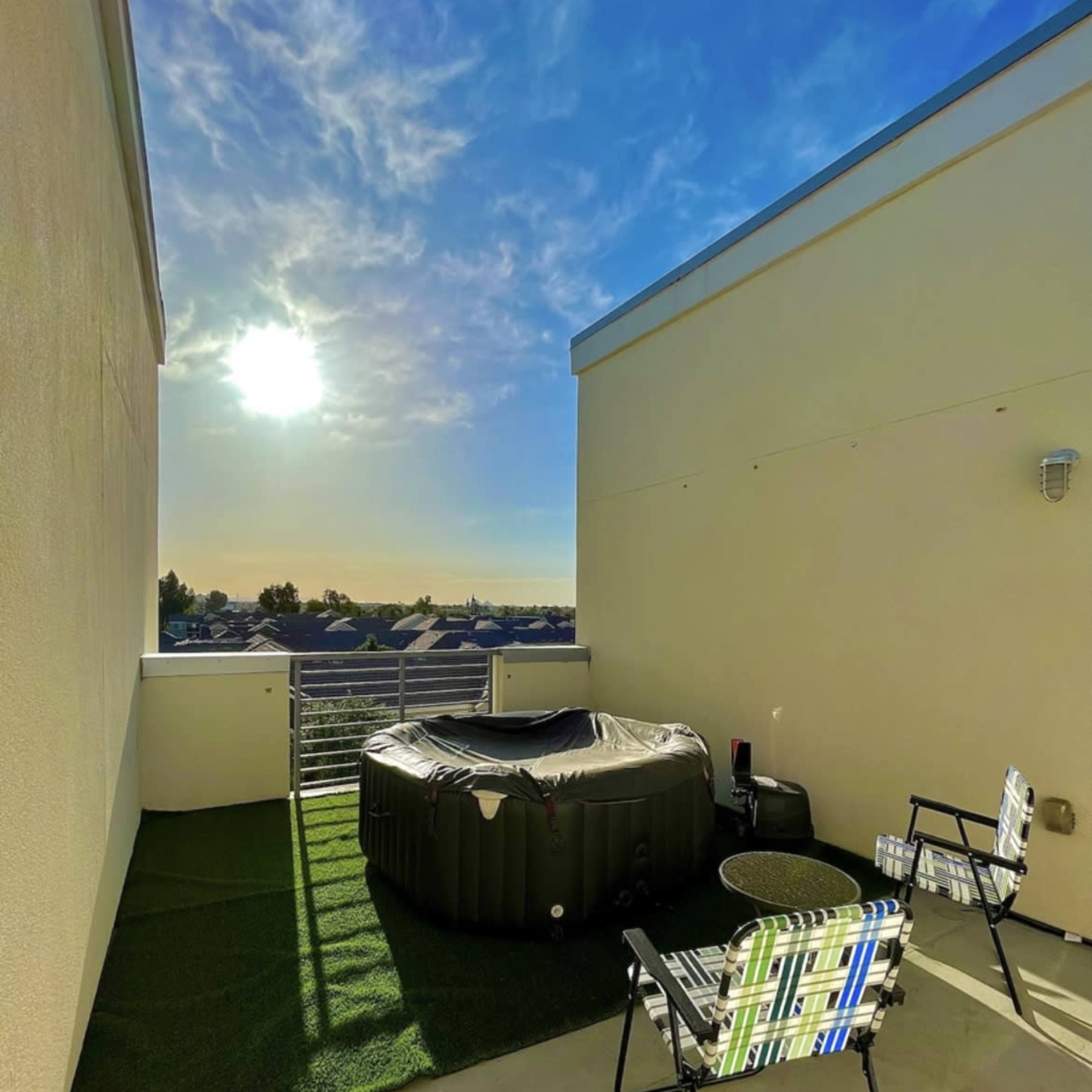 A hot tub is situated on a rooftop terrace with two chairs, set against a backdrop of a clear sky and distant rooftops.