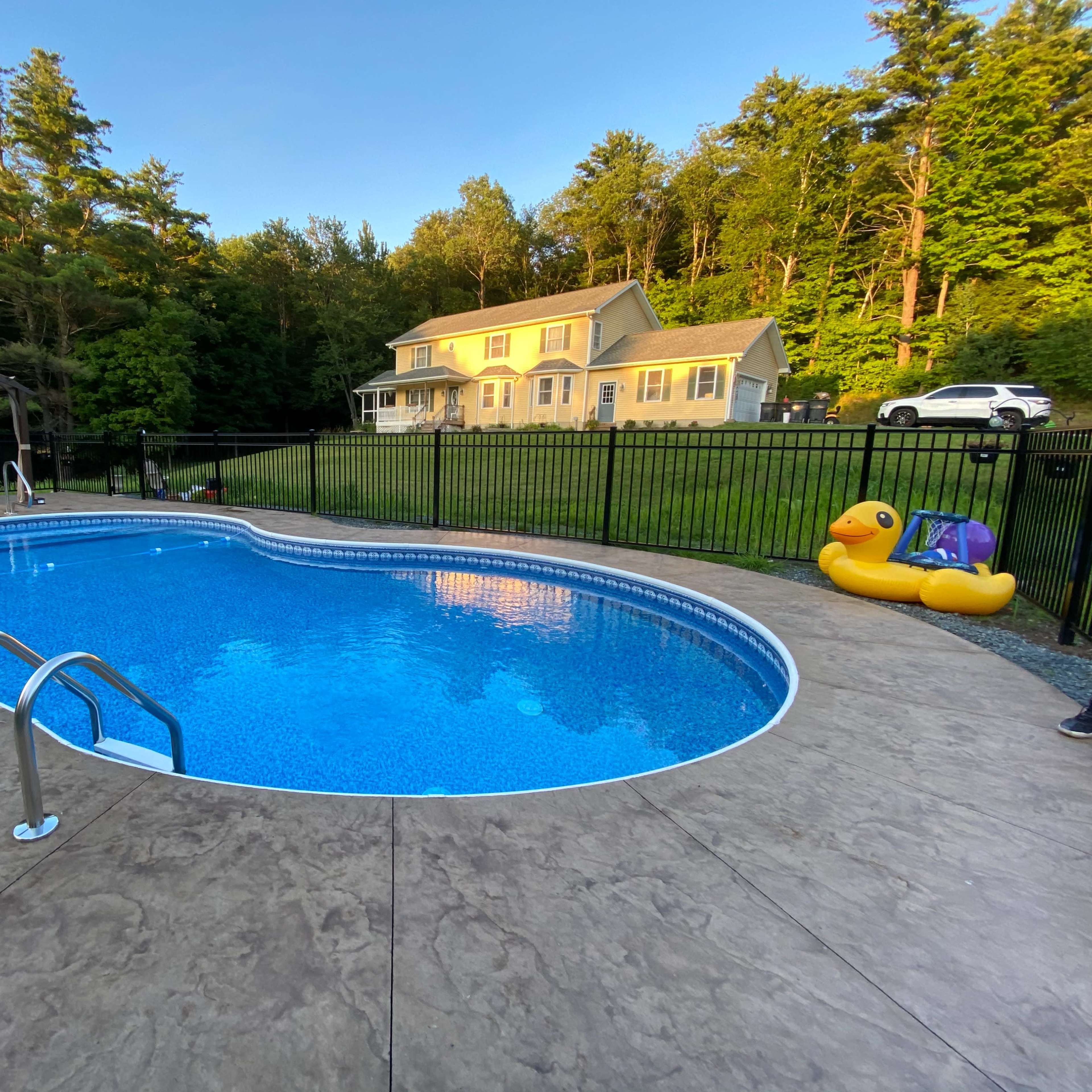 A blue swimming pool with ladders, surrounded by a concrete patio, features a yellow inflatable duck and a house in the background, all set against a wooded area.