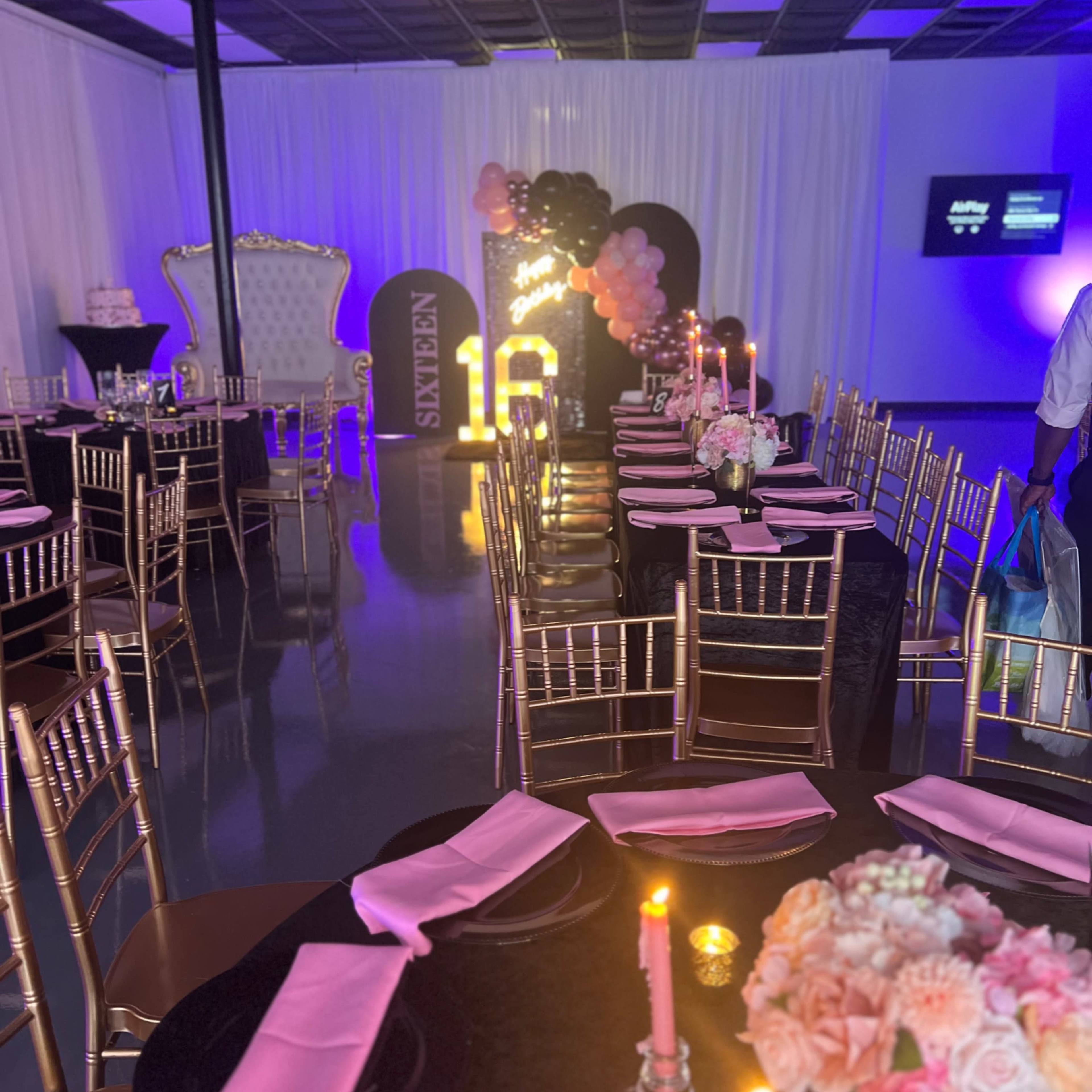 A party setup with elegantly arranged tables, gold chairs, and a decorative backdrop featuring the number "16" in lights, adorned with pink and black balloons.