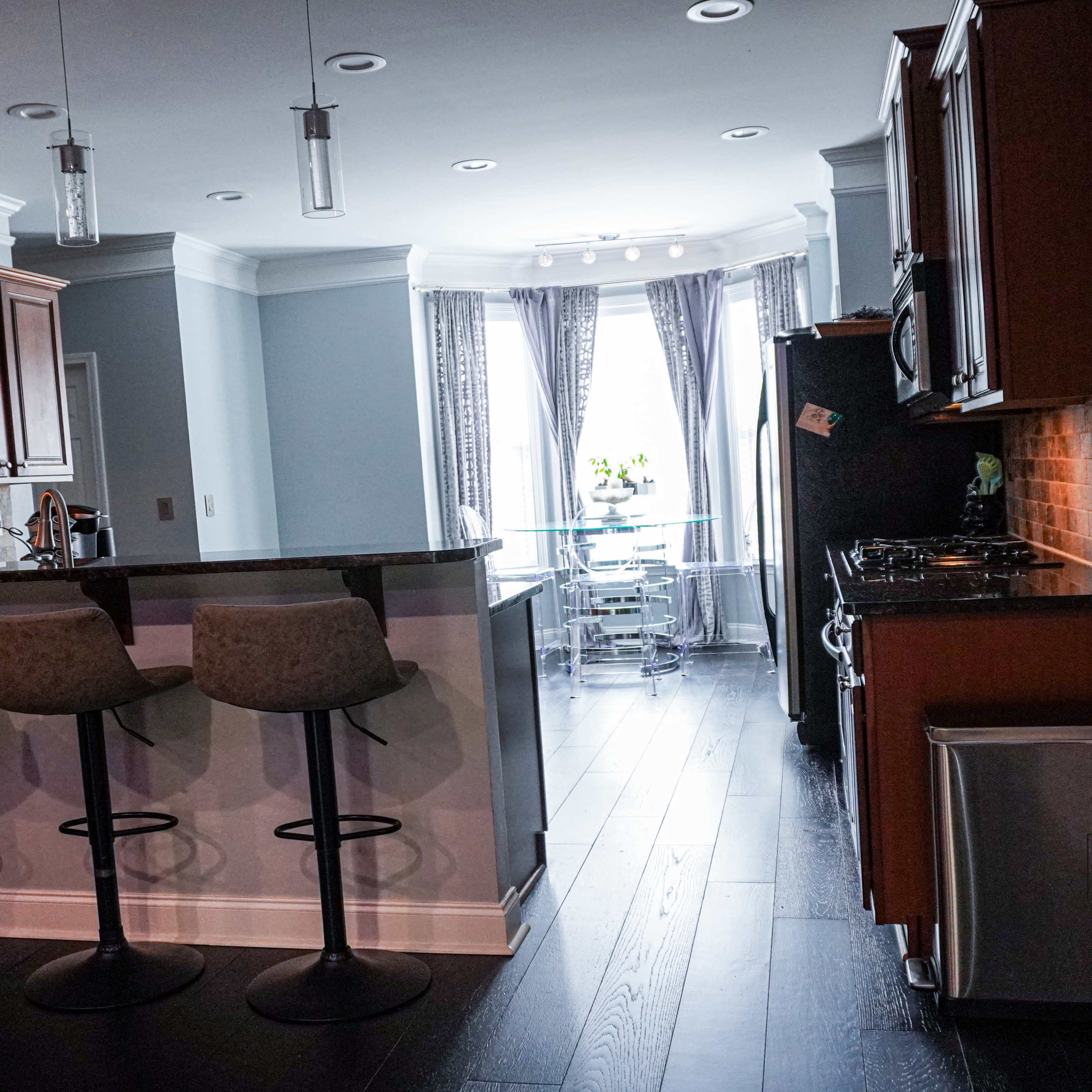 A modern kitchen features a bar with three stools, wooden cabinetry, and stainless steel appliances, leading to a dining area with large windows.