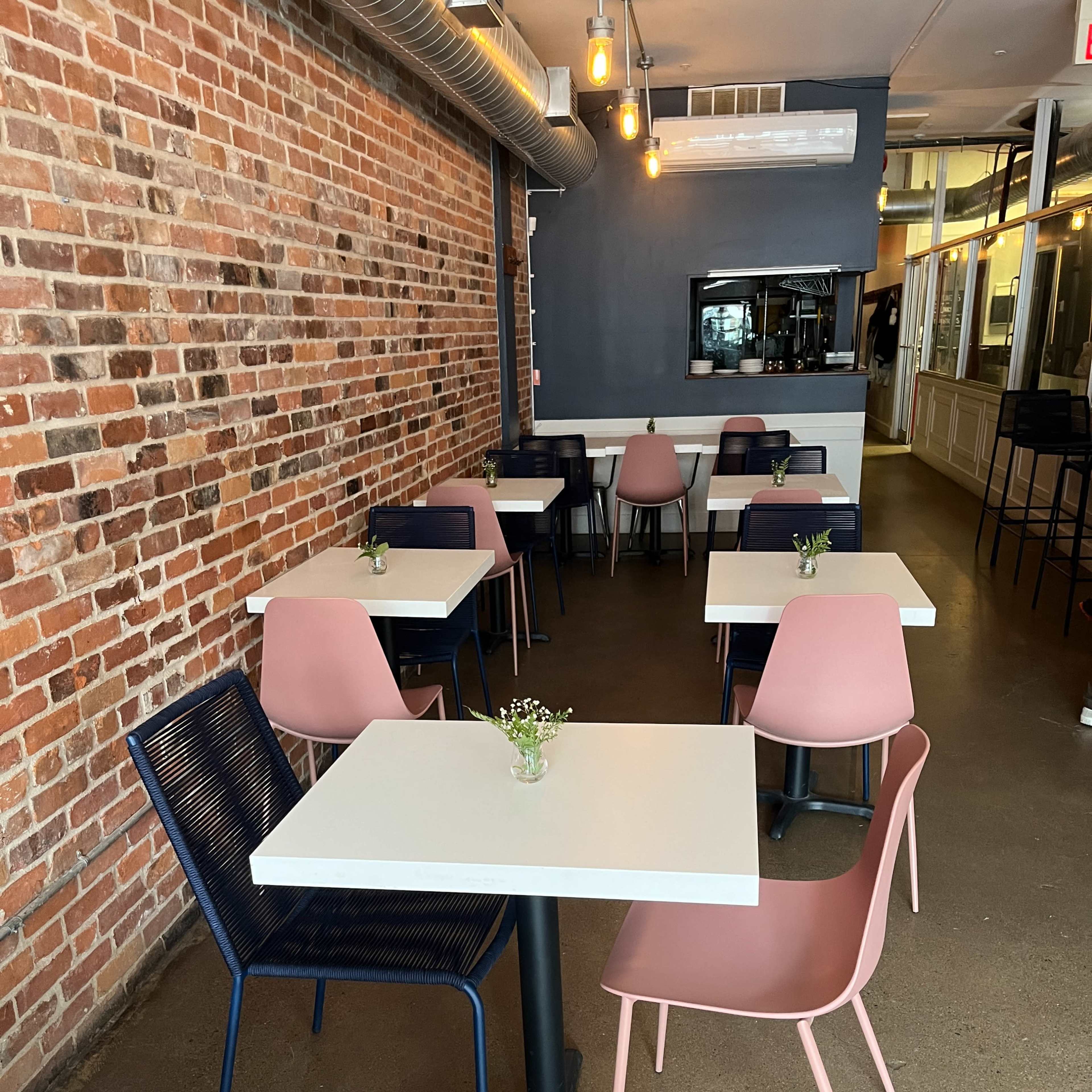 A modern restaurant interior featuring several tables with white tops and pink and navy chairs, along a brick wall with pendant lights above.
