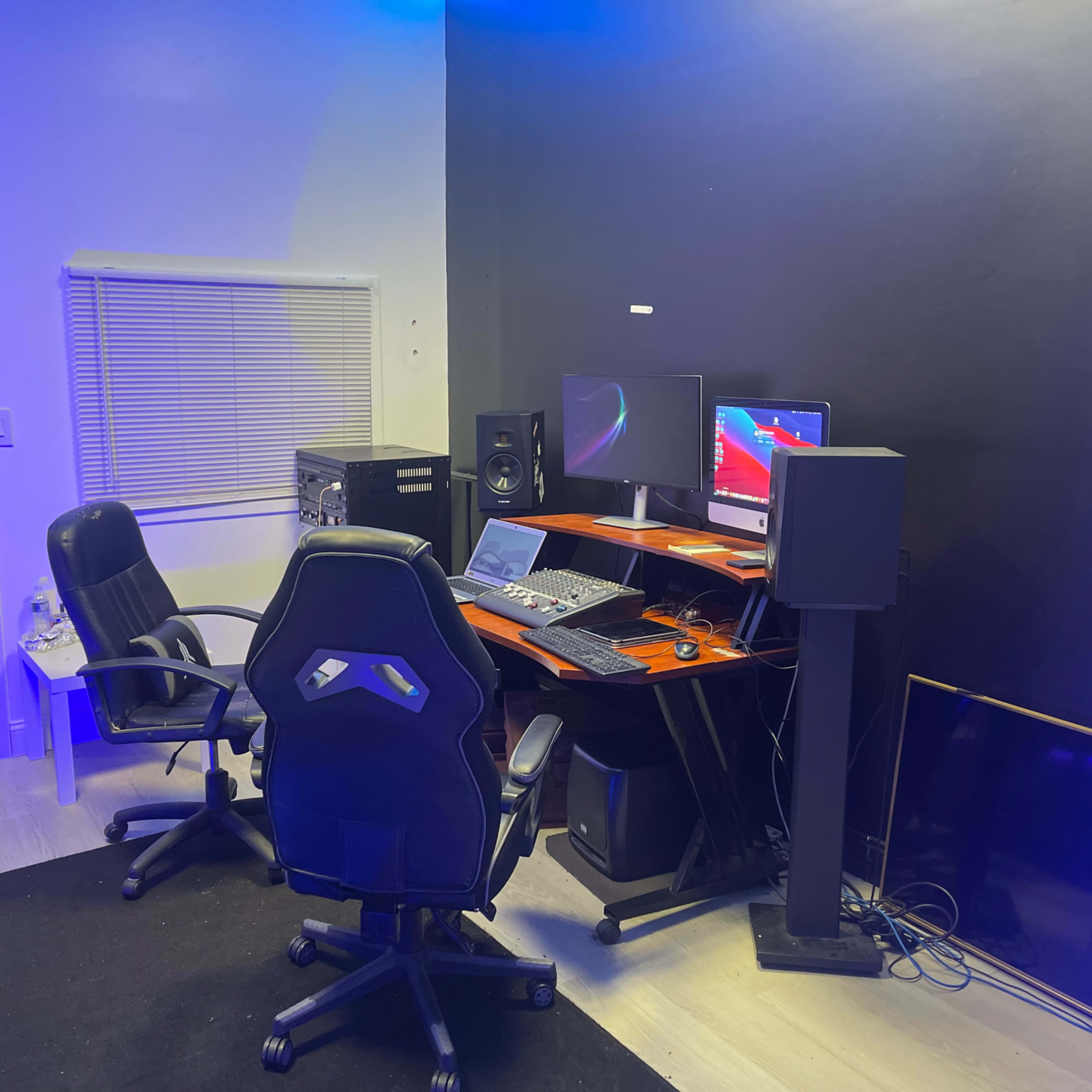 A modern home studio setup with two black chairs, a desk containing audio equipment, two monitors, and speakers against a partially illuminated dark wall.