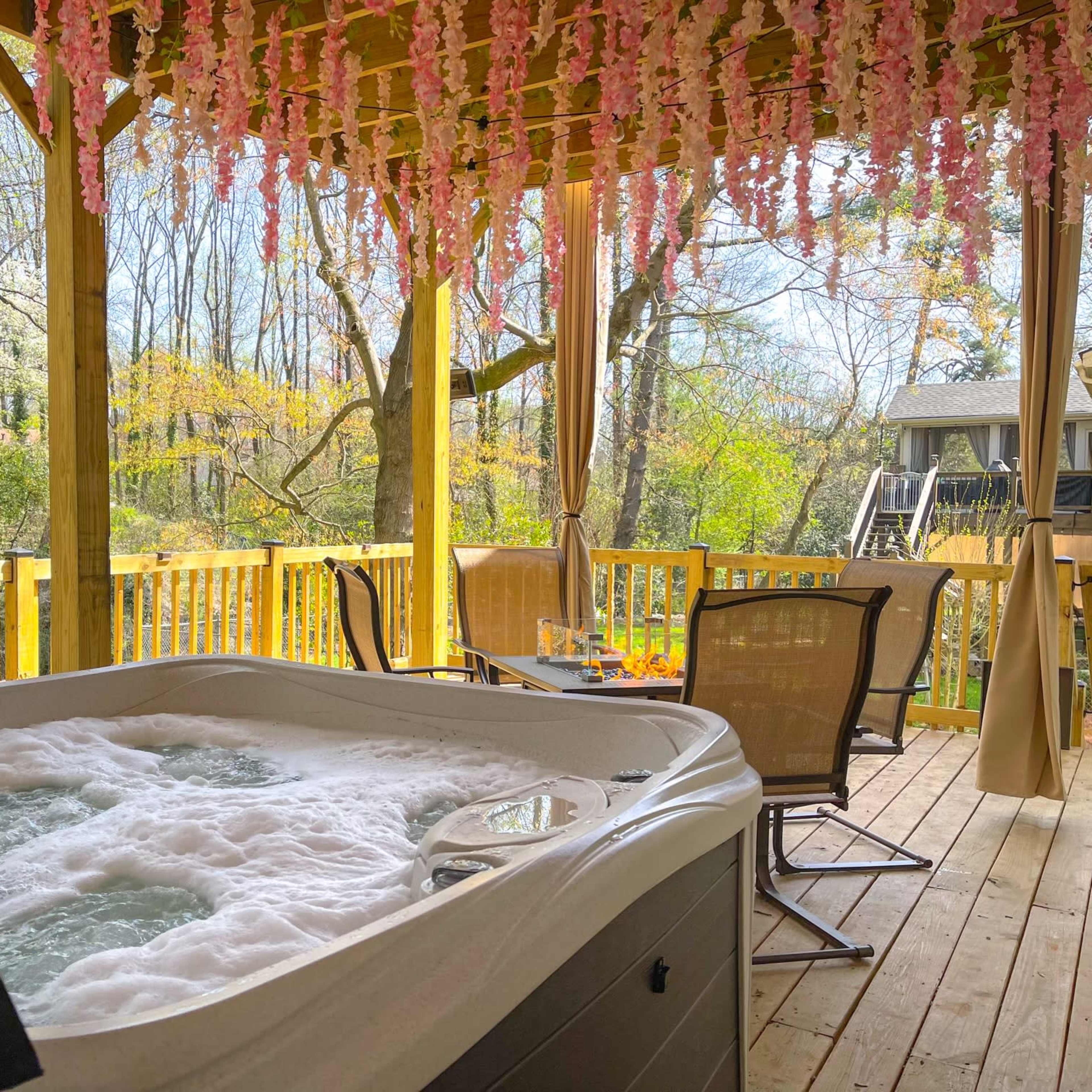 A hot tub with bubbling water is situated under a wooden deck adorned with pink flowers, alongside outdoor seating and a view of trees.