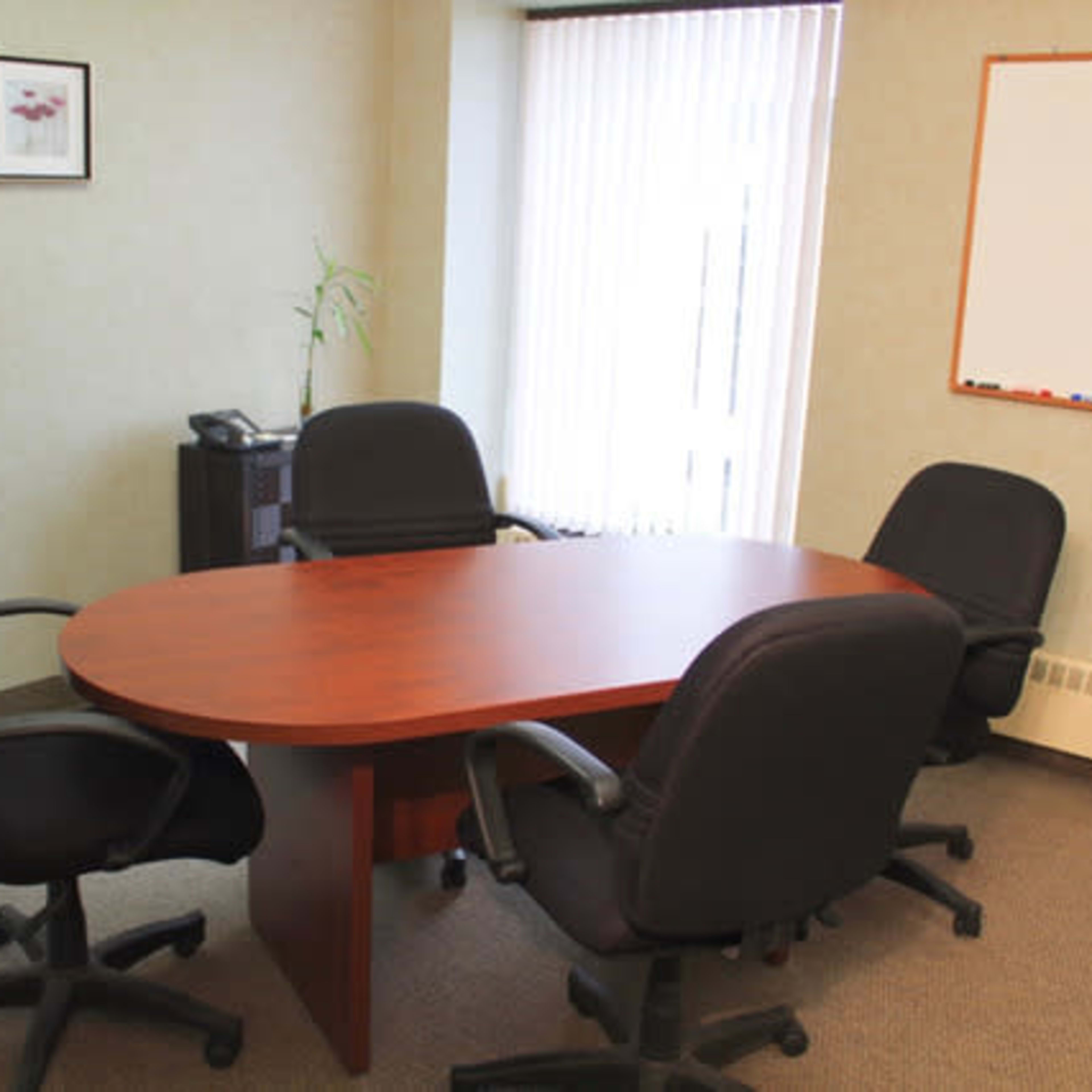 A conference room features a wooden oval table surrounded by four black office chairs, with a whiteboard and two framed pictures on the wall.