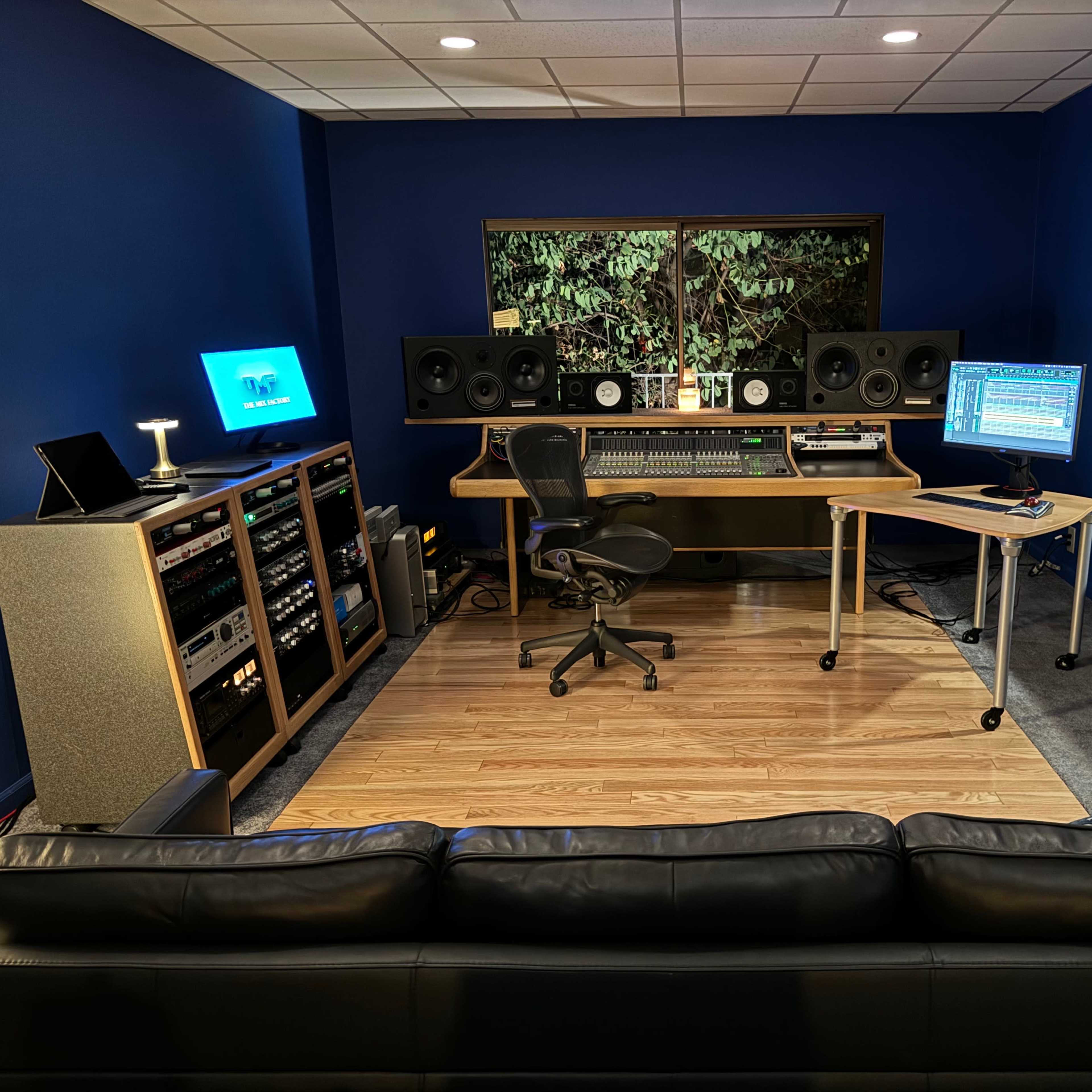 A modern music studio featuring a control desk with audio equipment, two monitors, speakers, and a couch in front of the workspace.