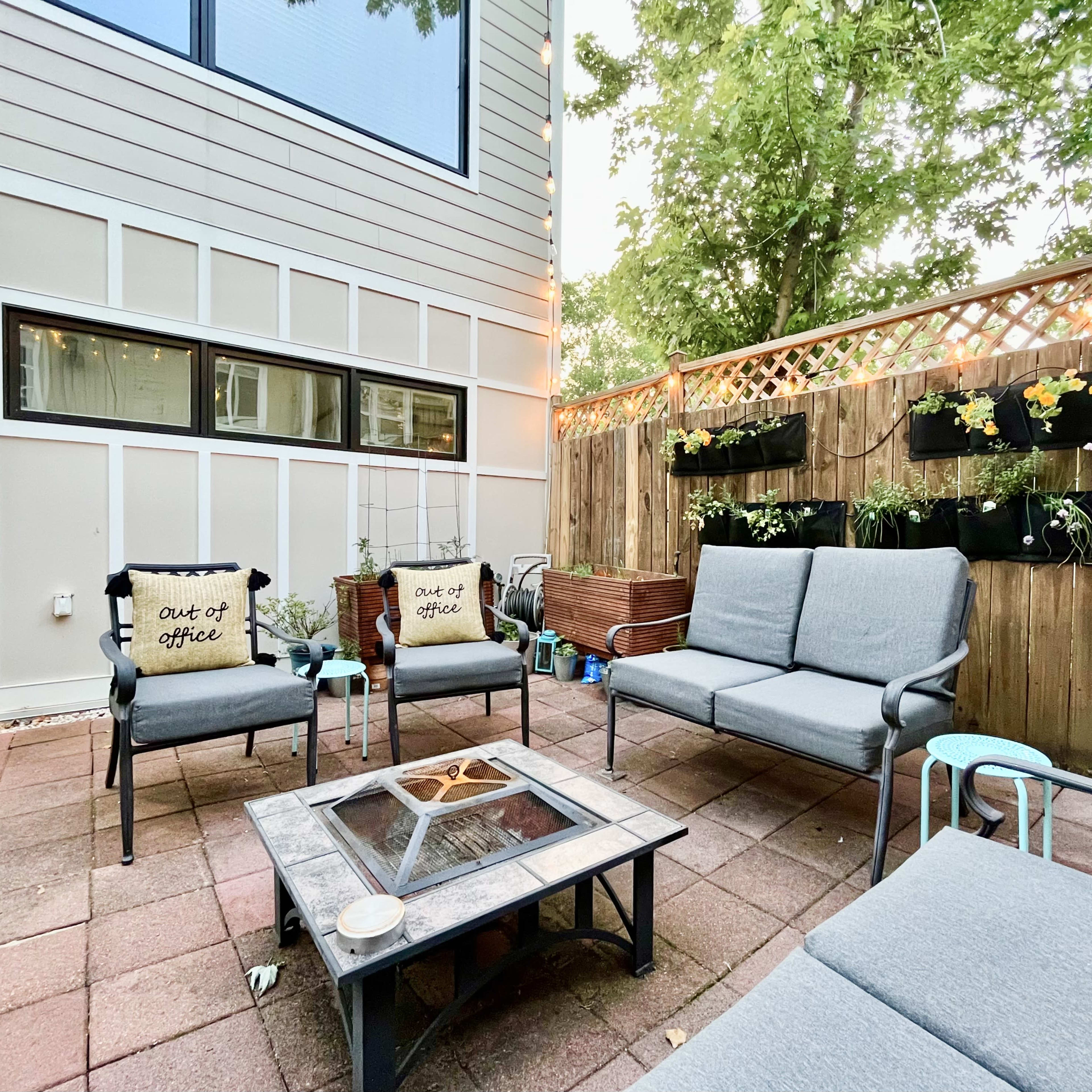 A patio area features a seating arrangement with gray cushions, a fire pit in the center, and potted plants along the fence.