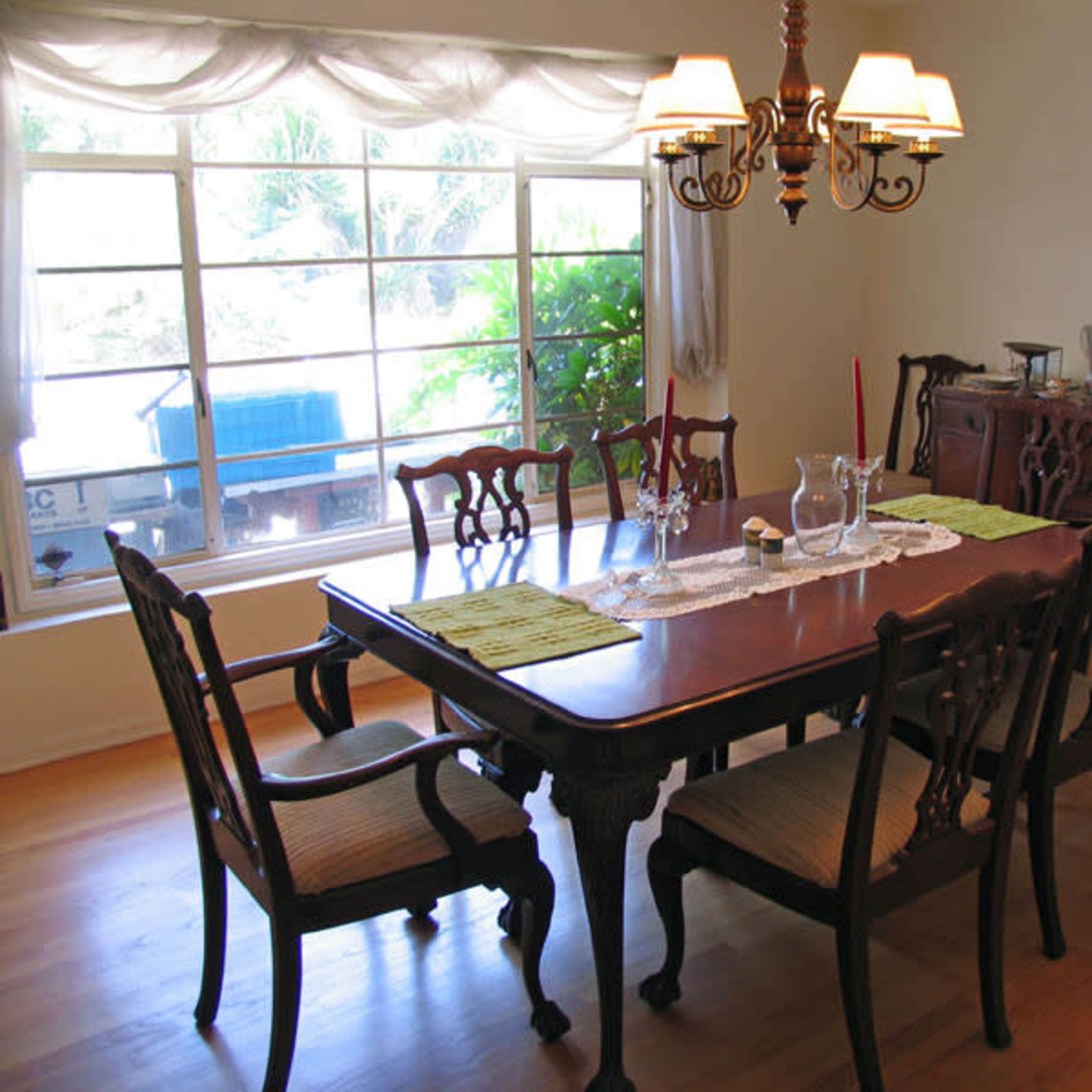 A dining room features a long wooden table surrounded by six chairs, with a chandelier above and a window overlooking greenery.