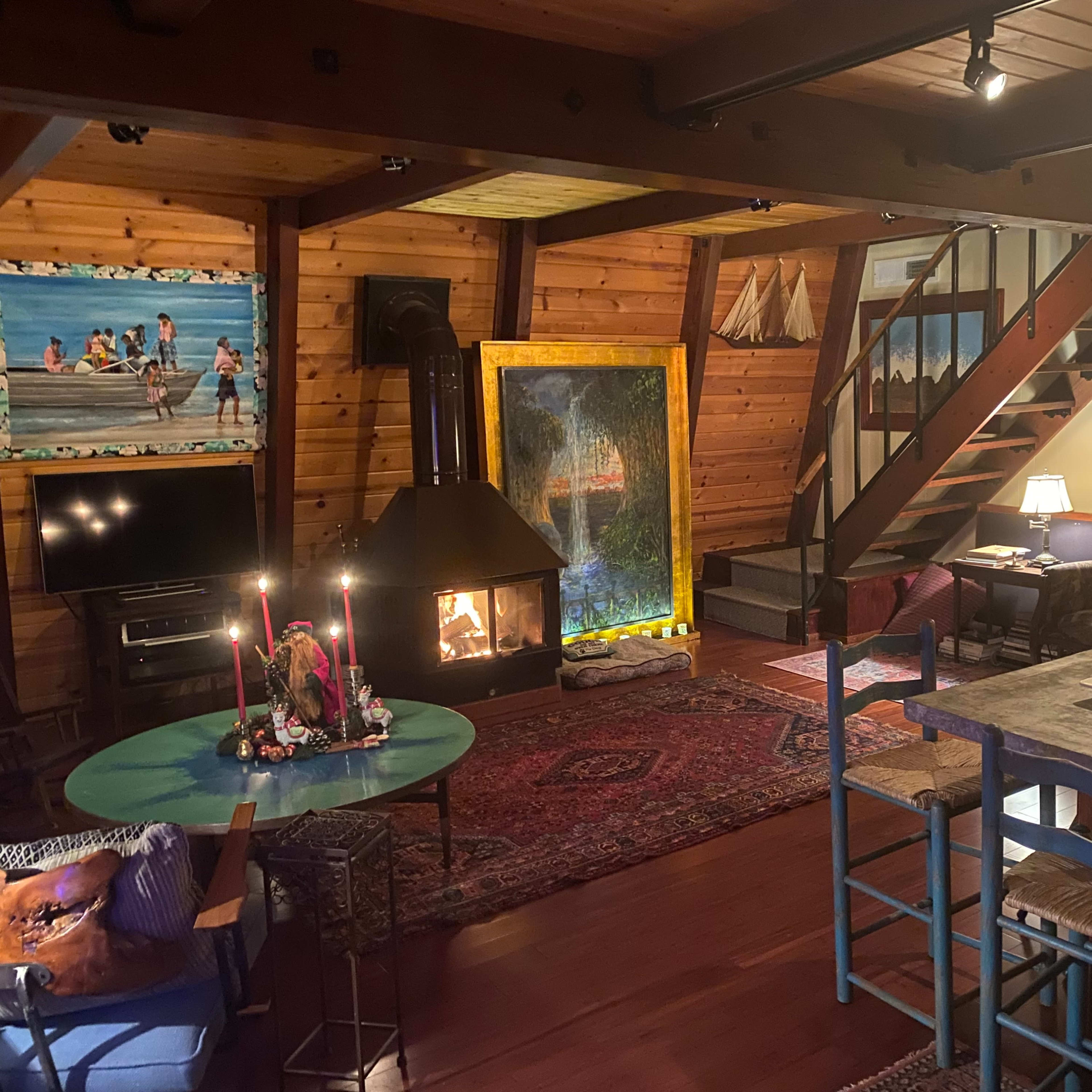 A cozy interior of a wooden cabin featuring a fireplace, a dining table, stairs, various artworks, and comfortable seating arrangements.