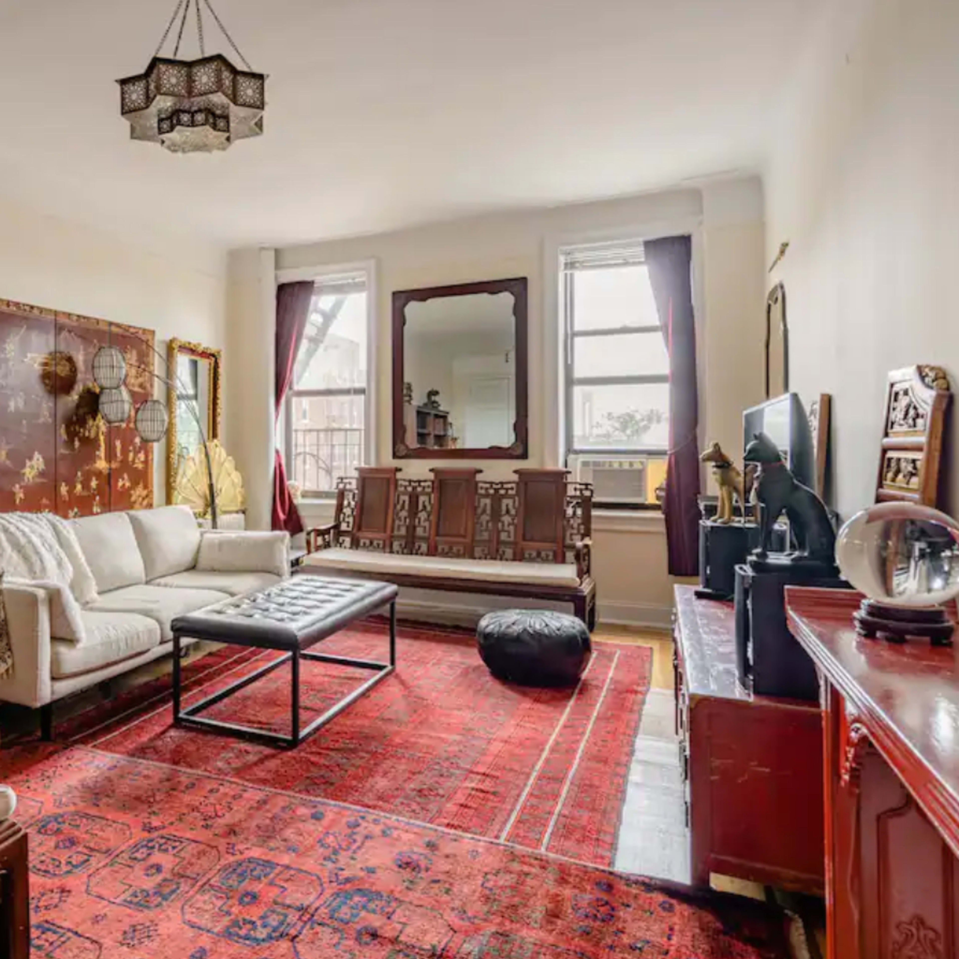 A living room featuring a white sofa, an ornate rug, a wooden bookshelf, and large windows with natural light.