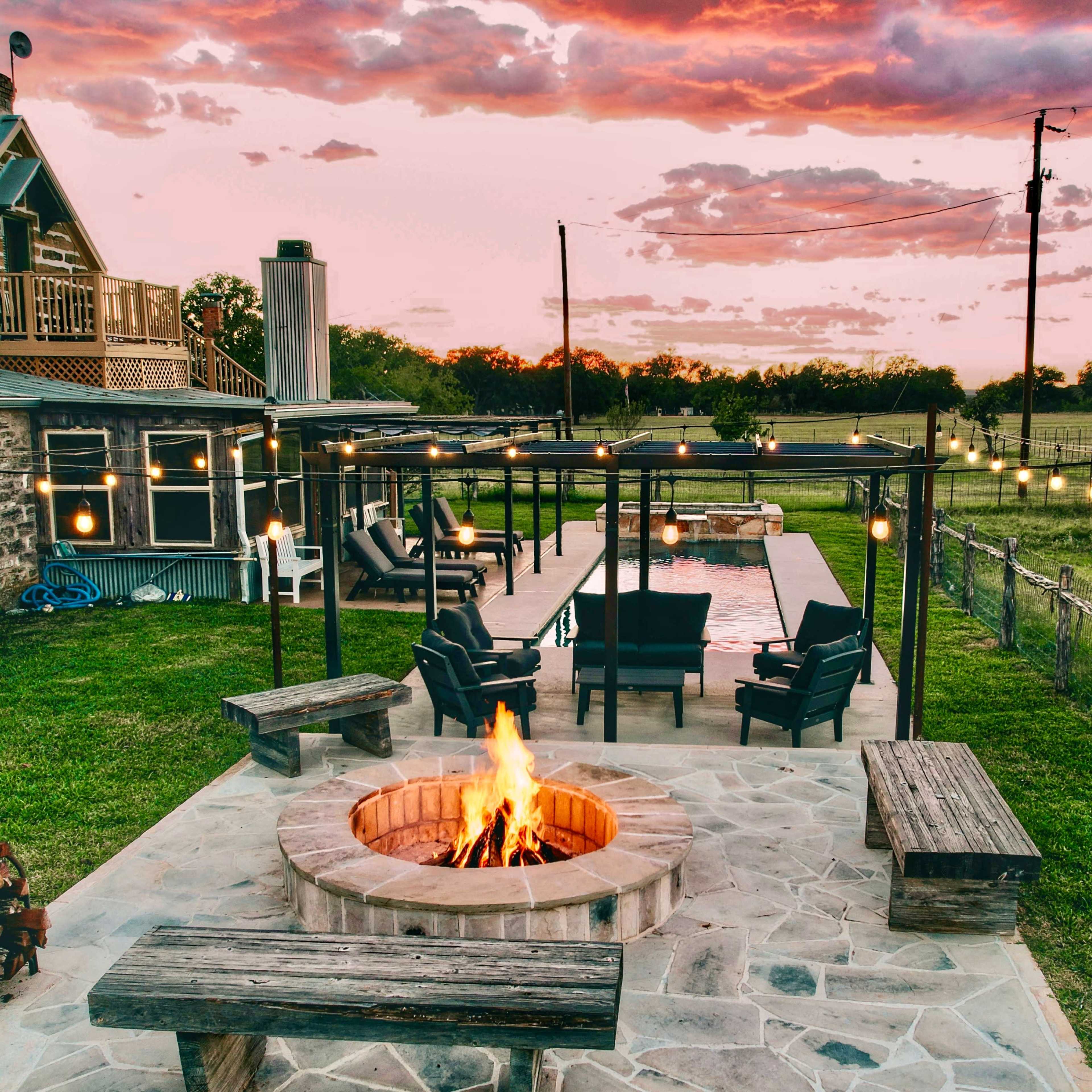A stone fire pit with benches surrounds a glowing fire, situated in a backyard with a pool and a house in the background under a colorful sunset sky.