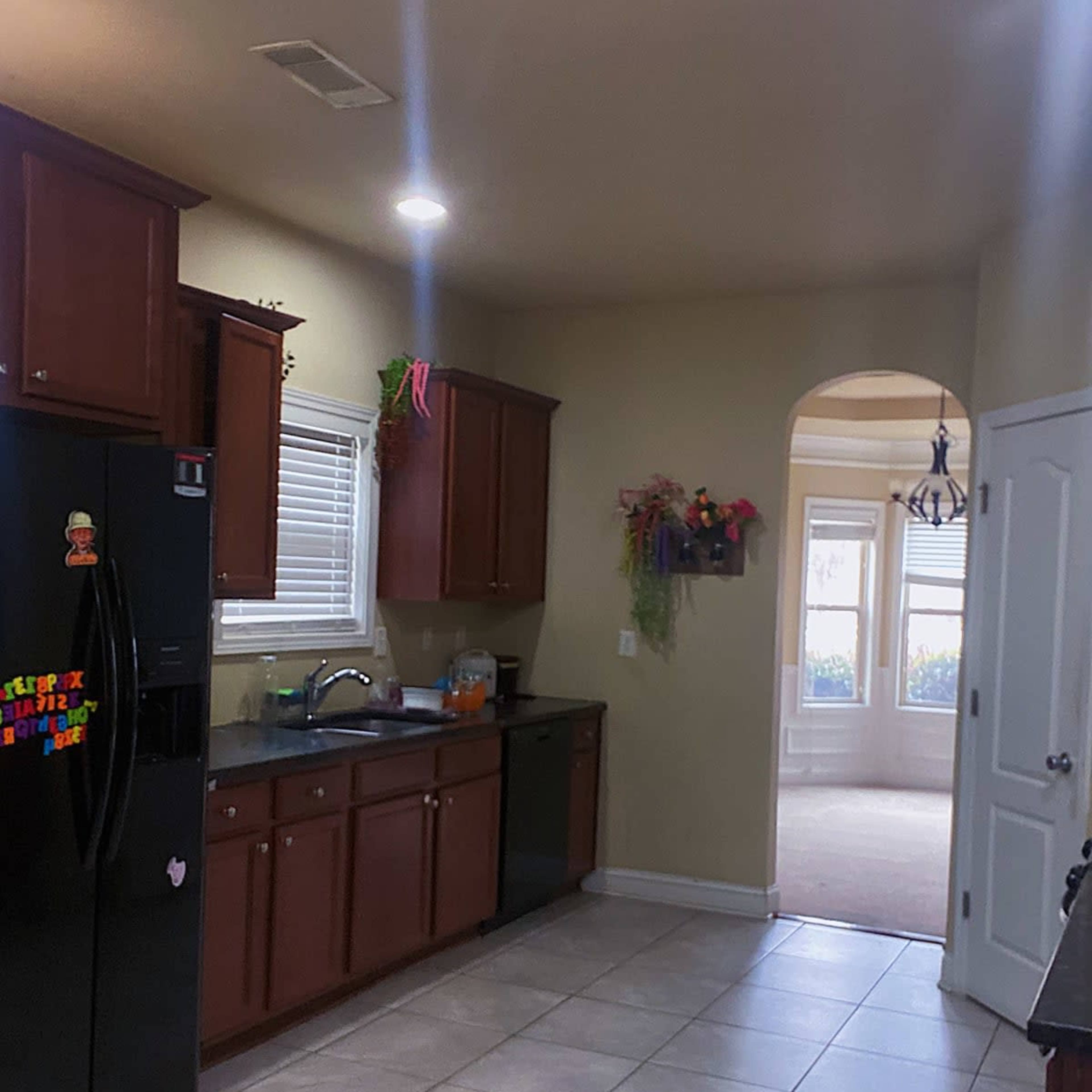 A kitchen with dark wooden cabinets, a black refrigerator, and tile flooring, leading to an arched doorway that opens into a carpeted room.