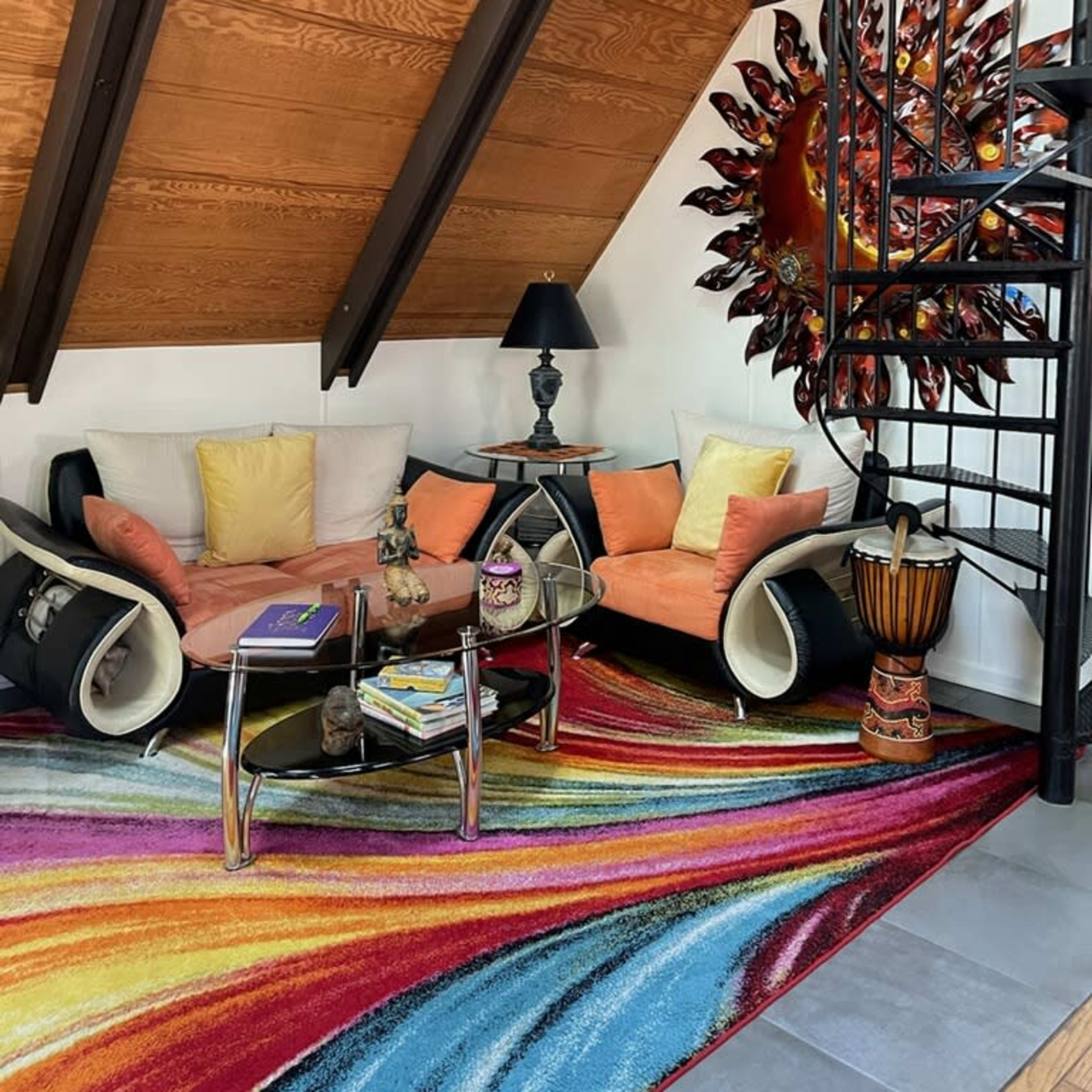 A cozy living room with a colorful area rug, a circular glass coffee table, two orange sofas, and an ornate wall decoration, all under a slanted wooden ceiling.