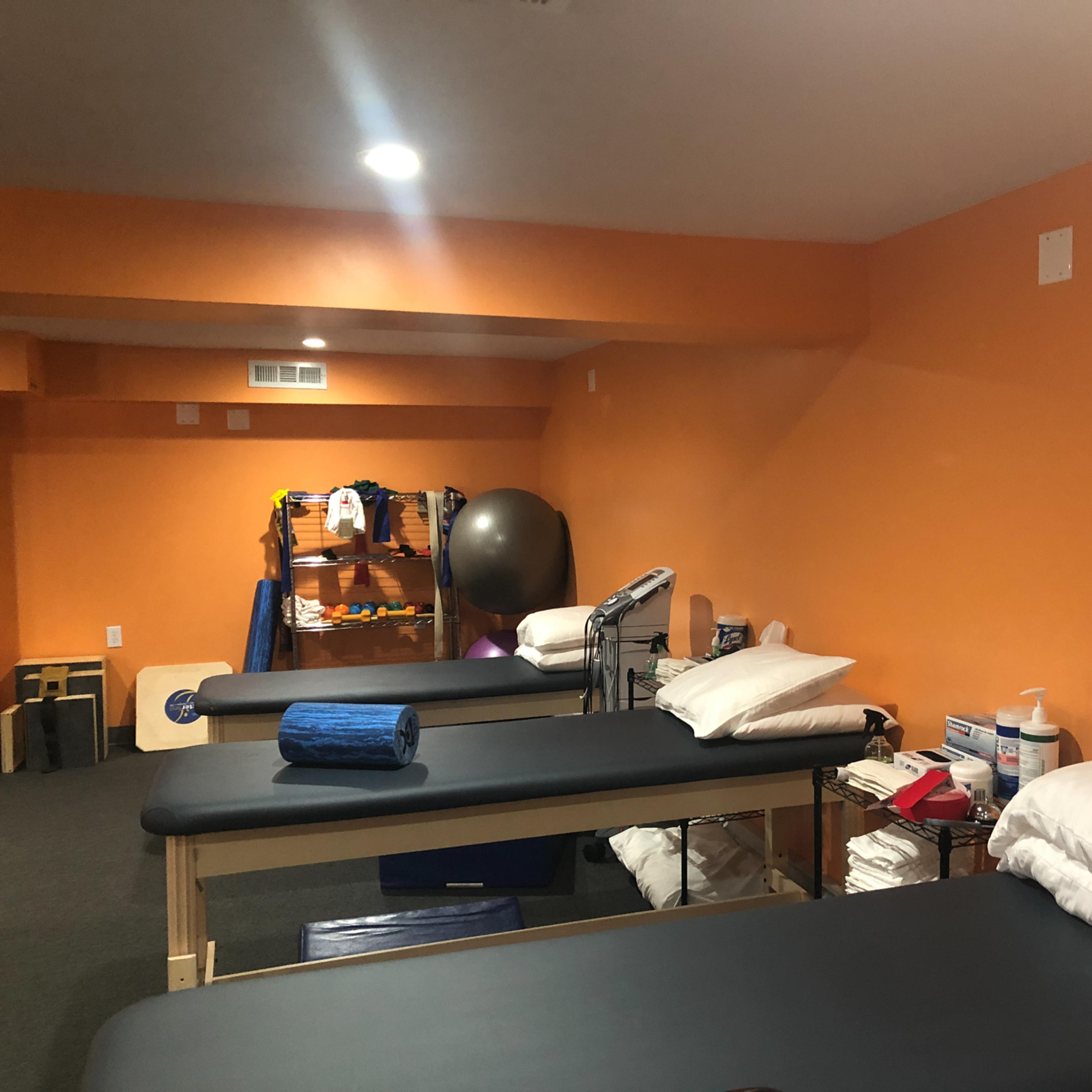 A room with several massage tables, orange walls, and various exercise equipment in a therapy or fitness setting.