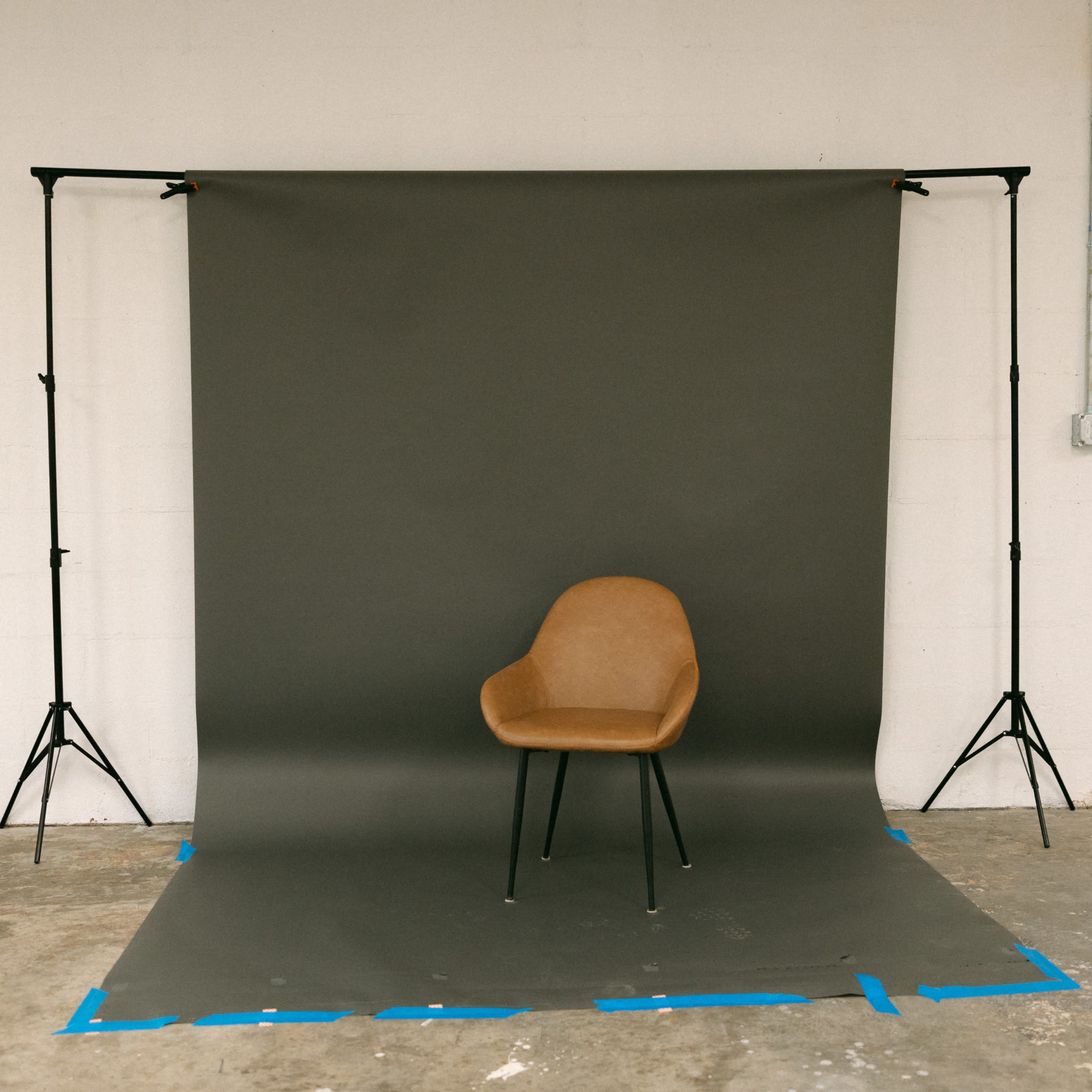 A brown chair is positioned on a gray backdrop supported by black stands in an empty room.