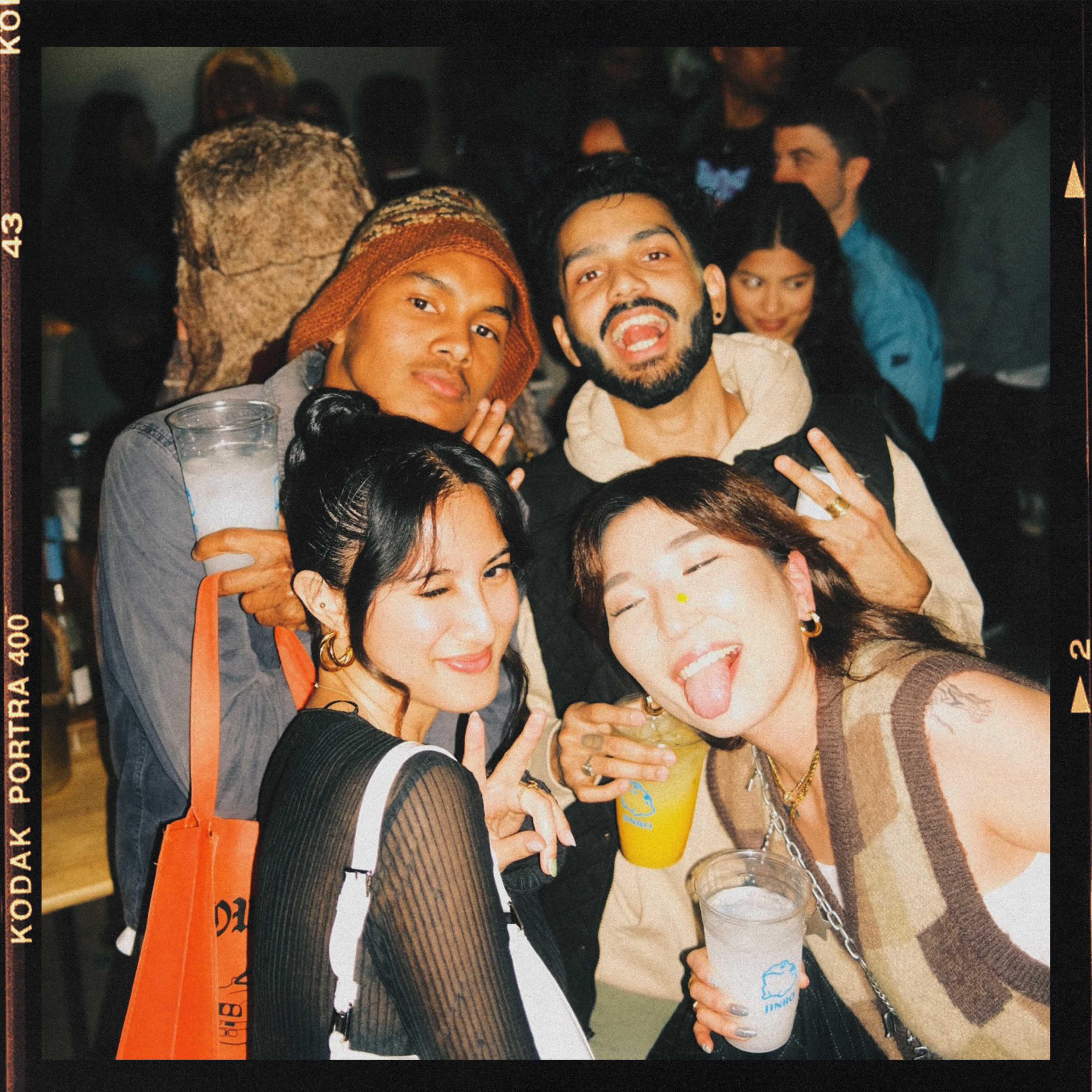 A group of four friends poses for a photo in a lively indoor setting, holding drinks and smiling at the camera.