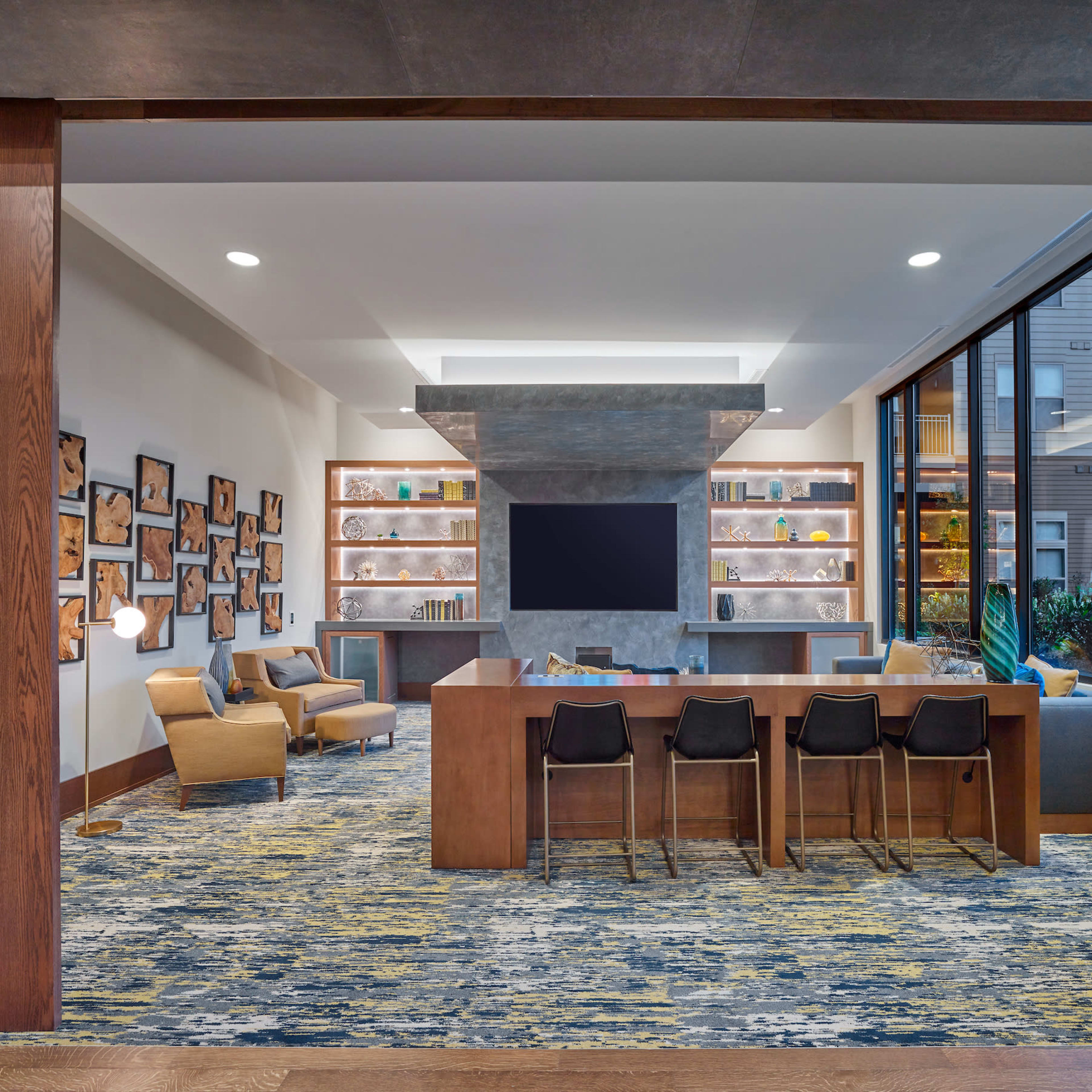 A modern lounge area featuring a large meeting table, comfortable seating, wall shelves with decorative items, and a large screen mounted on the wall.