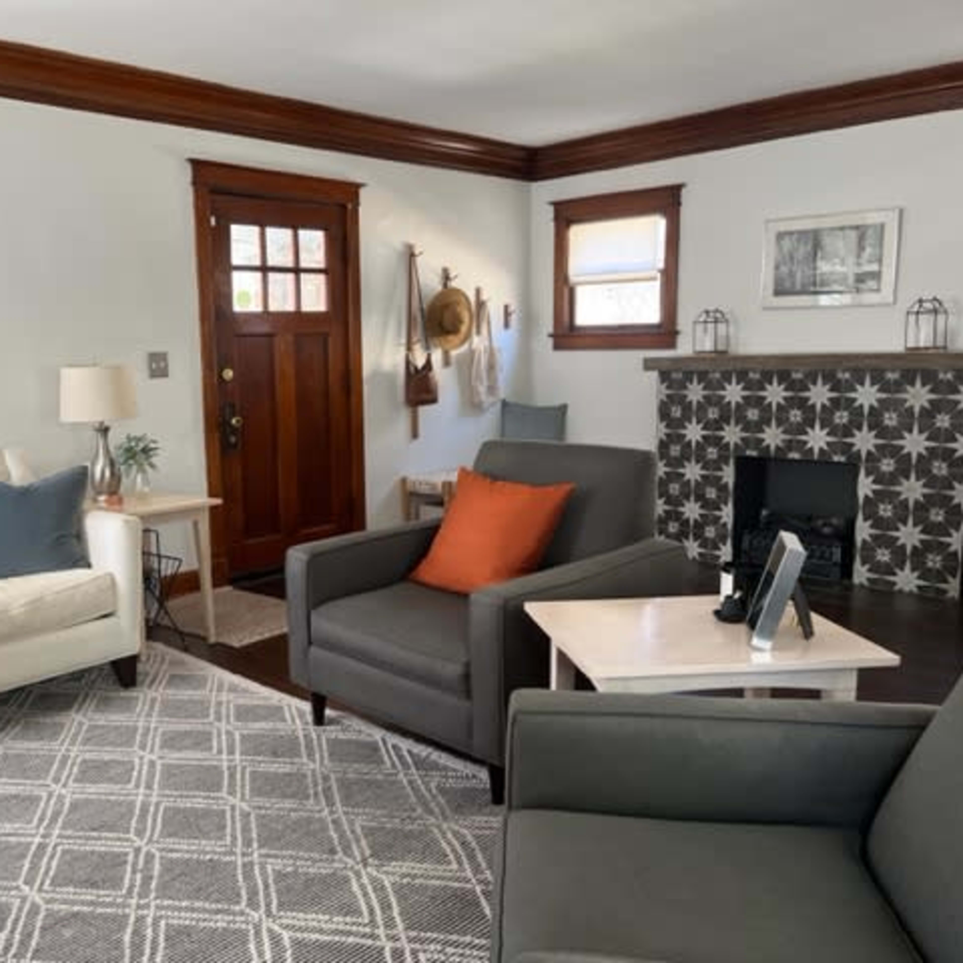 The living room features a light-colored sofa, two gray armchairs, a small side table, and a tiled fireplace, with wooden trim and large windows allowing natural light.