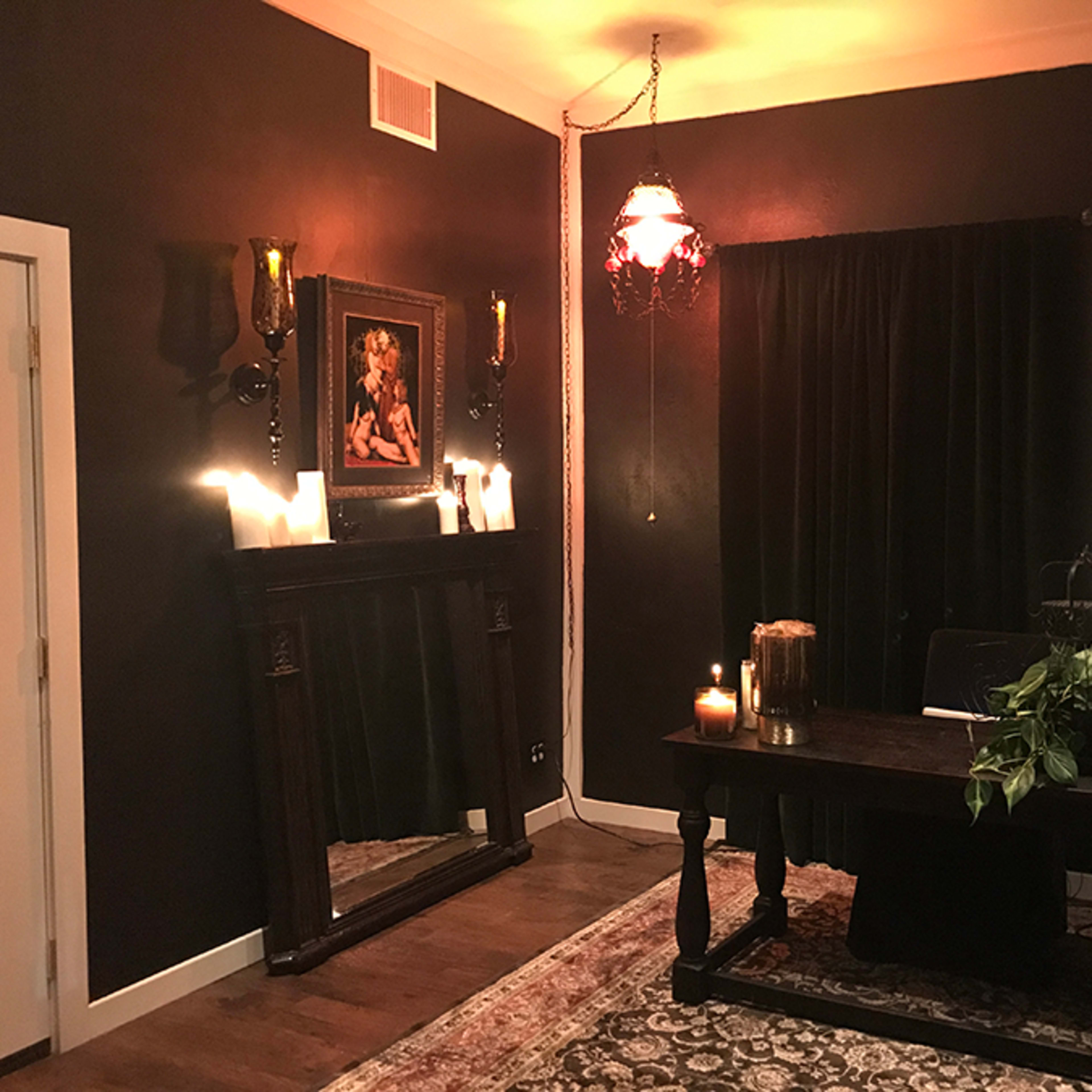 A dimly lit room with dark walls, featuring a wooden desk, a large mirror, candles, and a curtain draped over a window.