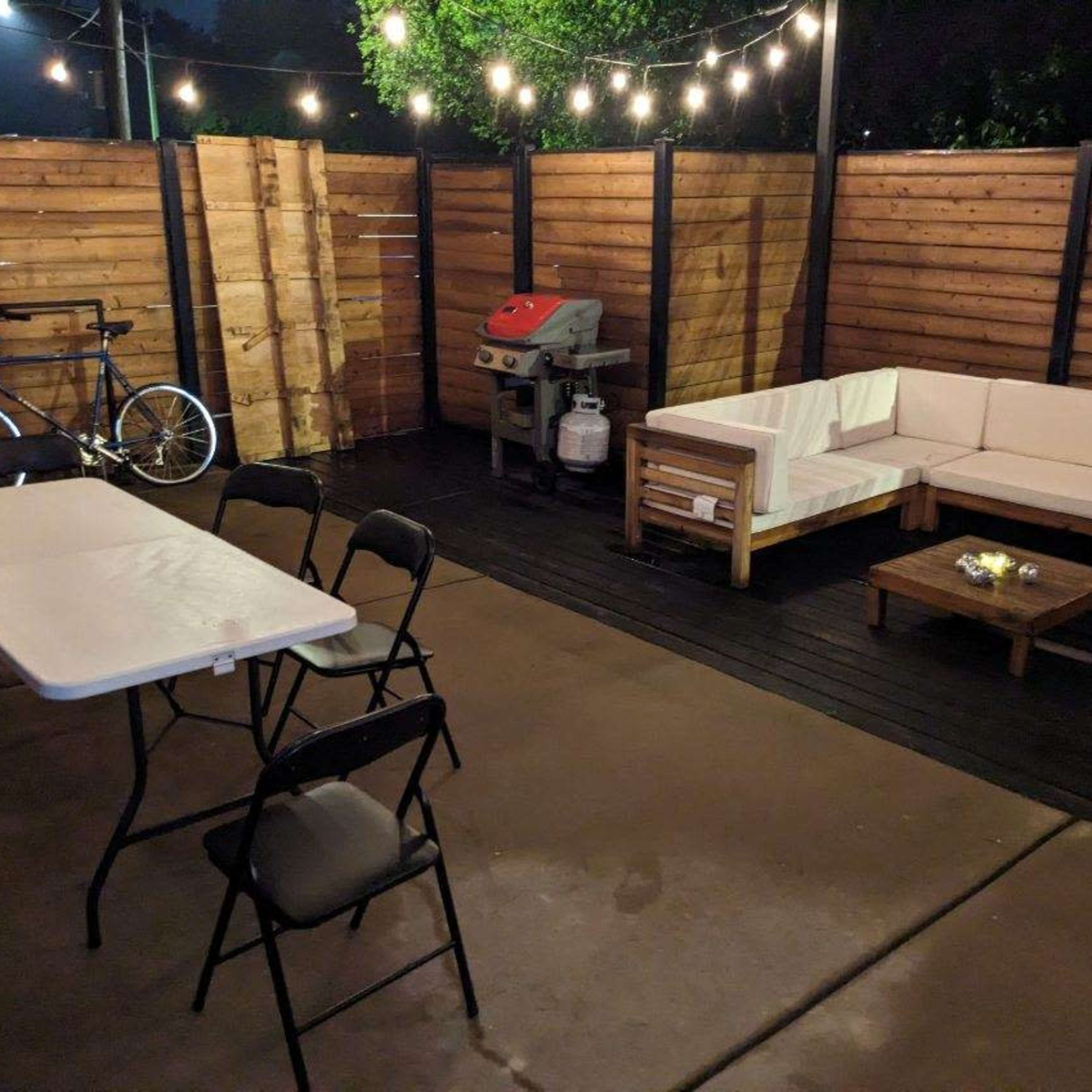 A patio area features a wooden couch set, a table with chairs, a grill, and a bike alongside a backdrop of wooden fences and string lights.
