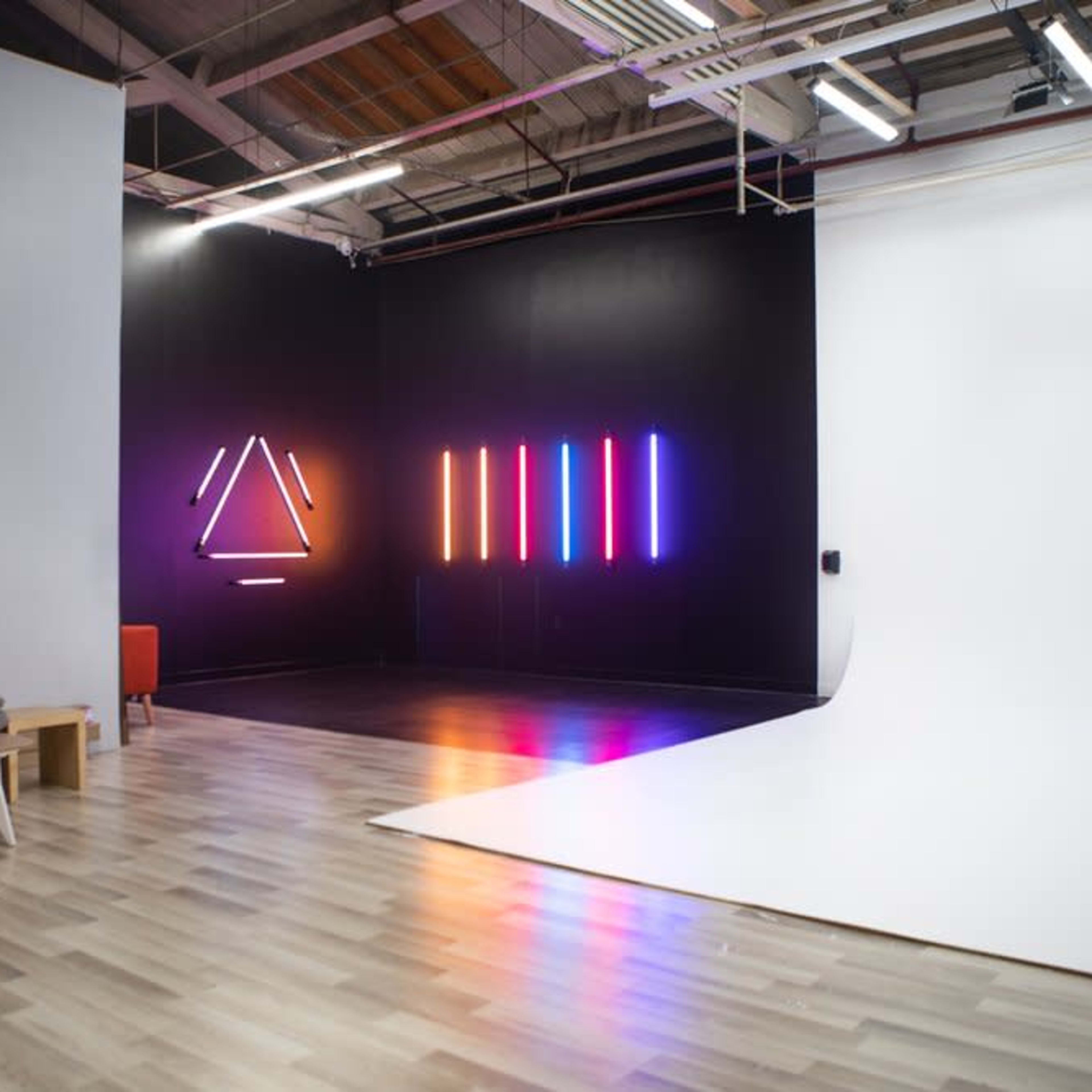 A modern studio space featuring a gray couch, colorful neon light displays on the black wall, and a large white backdrop area.